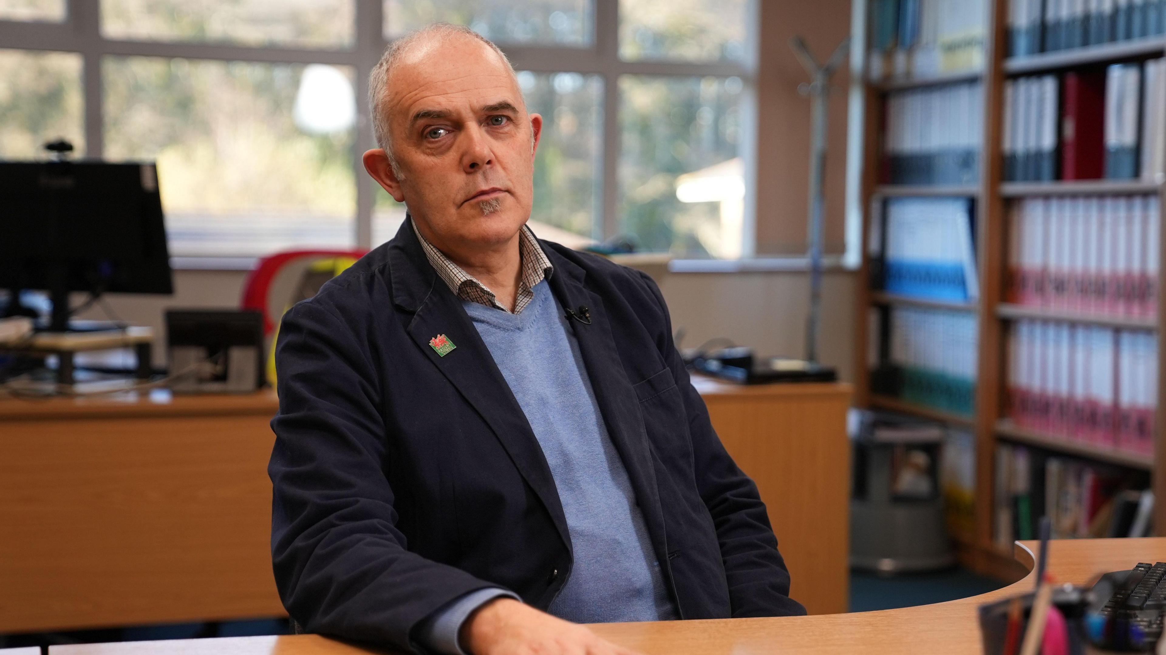 Sion Amlyn sits at his brown wood desk, he wears a shirt with a blue jumper overtop and a navy blazer. He has silver hair and a small bearded patch under his chin and stares blankly into the camera. 