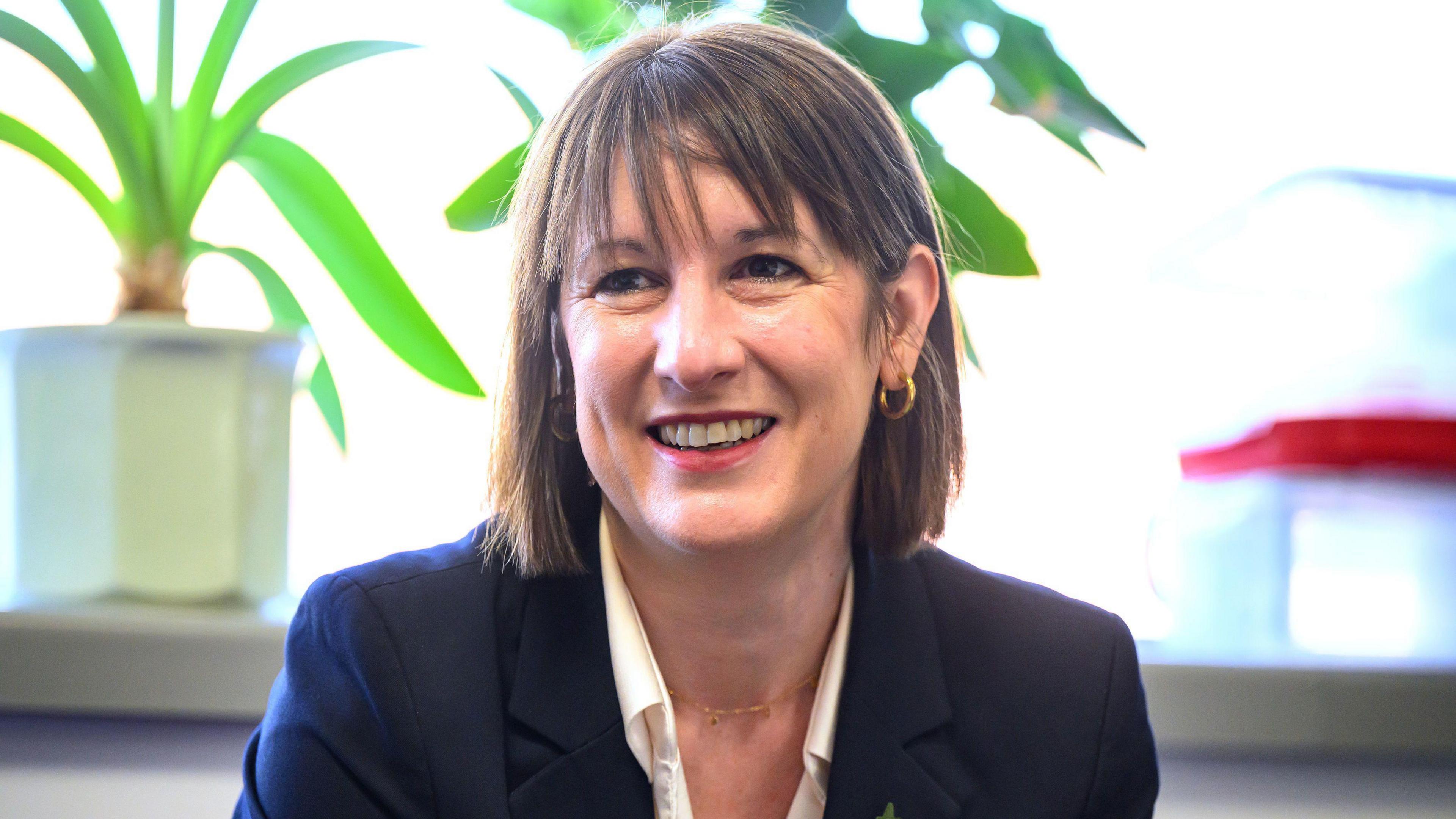 A woman in a navy blouse and white shirt looks off to the right with a plant pot over her right shoulder.