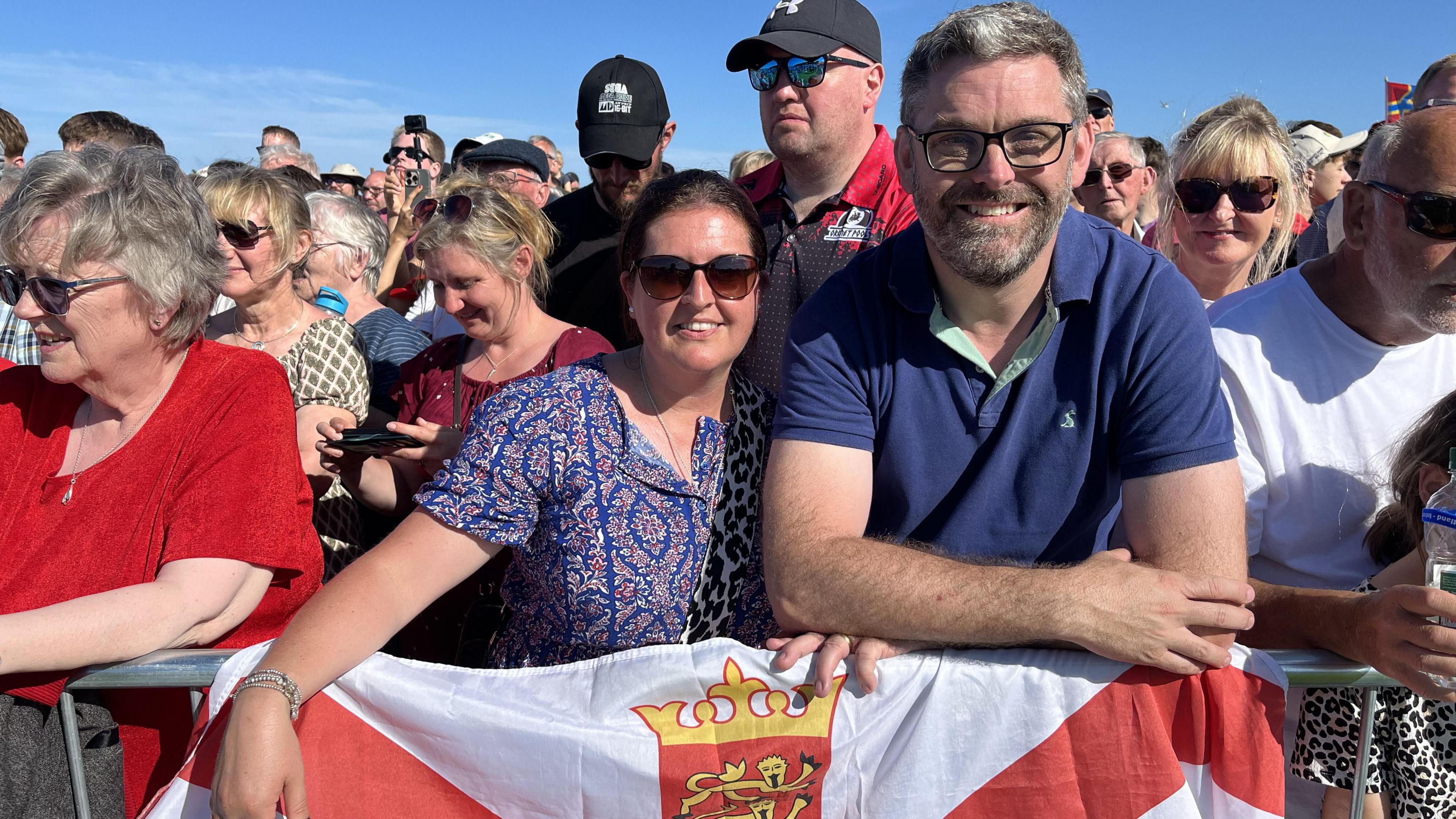 Lucy and Nick Le Boutillier are standing at the front of a crowd at the opening ceremony, leaning on a barrier with a Jersey flag.
