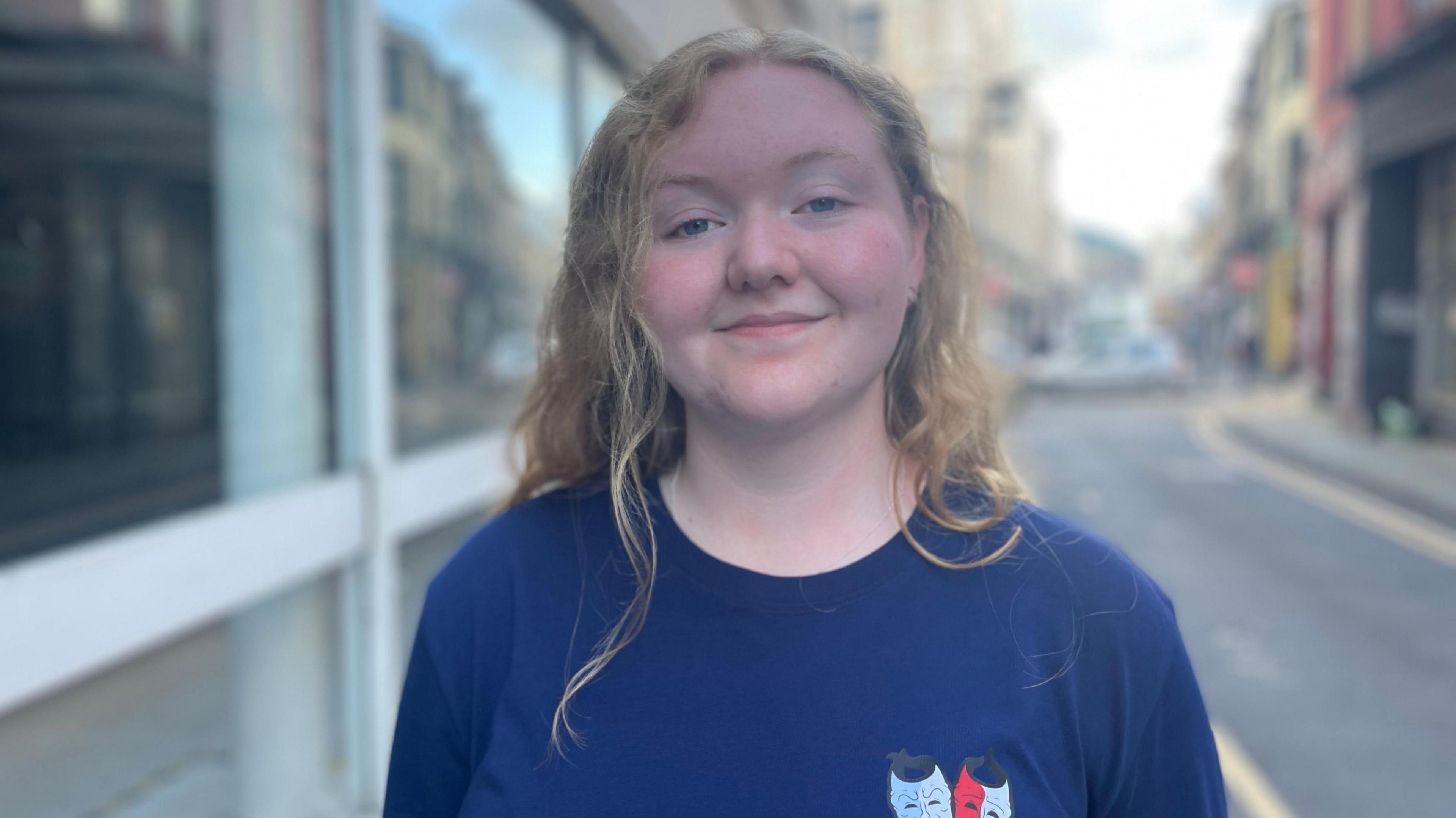 Megan, a young woman with long blonde wavy hair, wearing a blue T-shirt smiling at the camera with the street behind her. It is a head and shoulders shot of her.