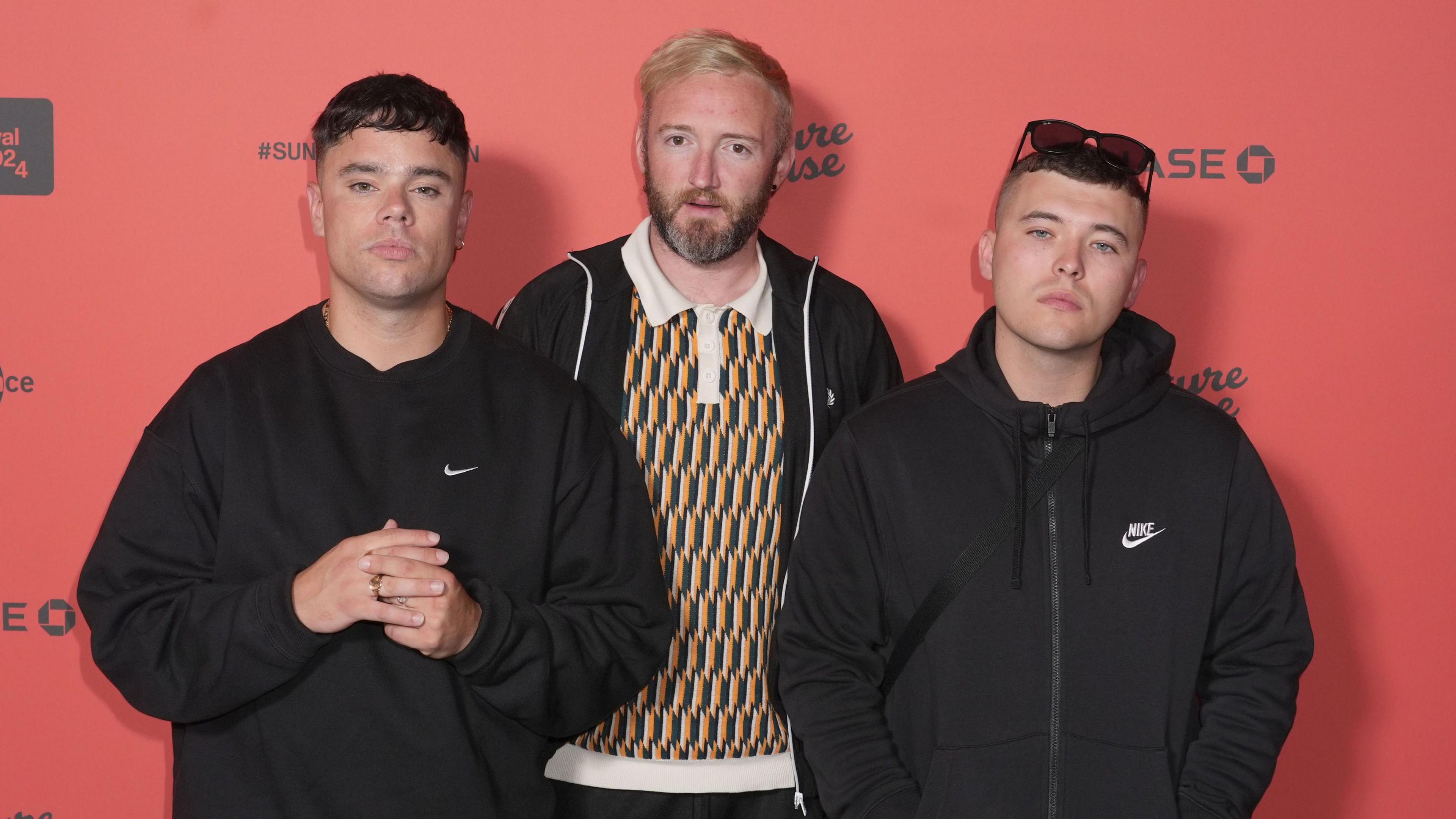 Kneecap members Moglai Bap, Mo Chara, DJ Provai standing together in front of a red wall.