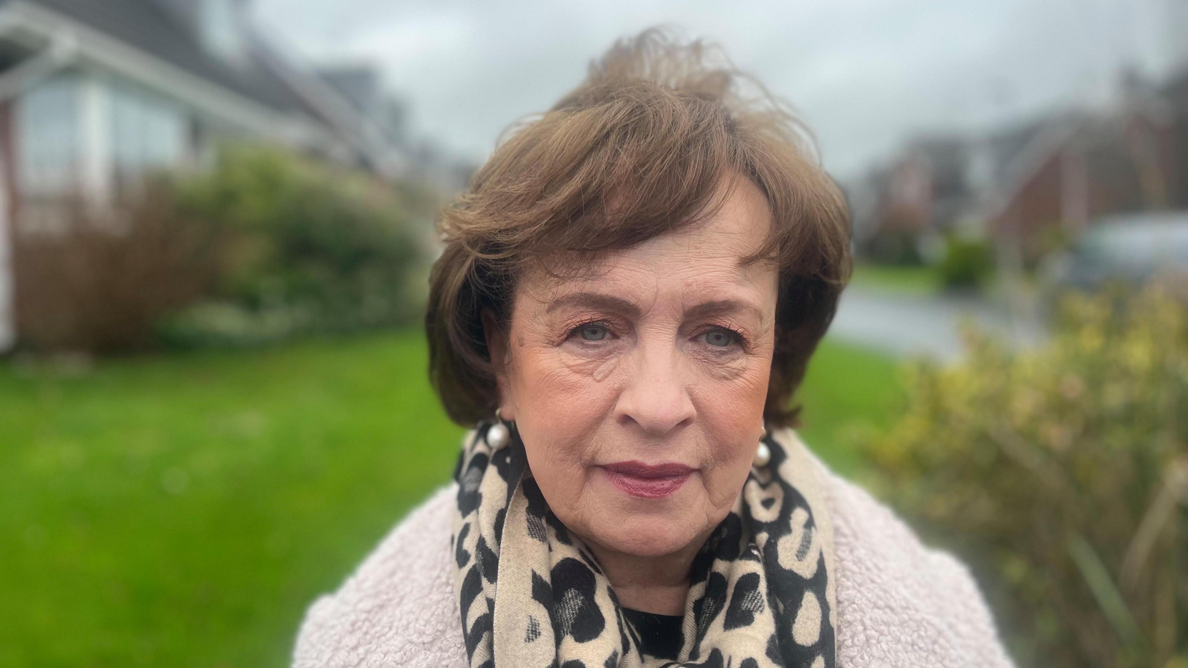 A woman with brown hair, animal print scarf and pink boucle cardigan stares into the camera