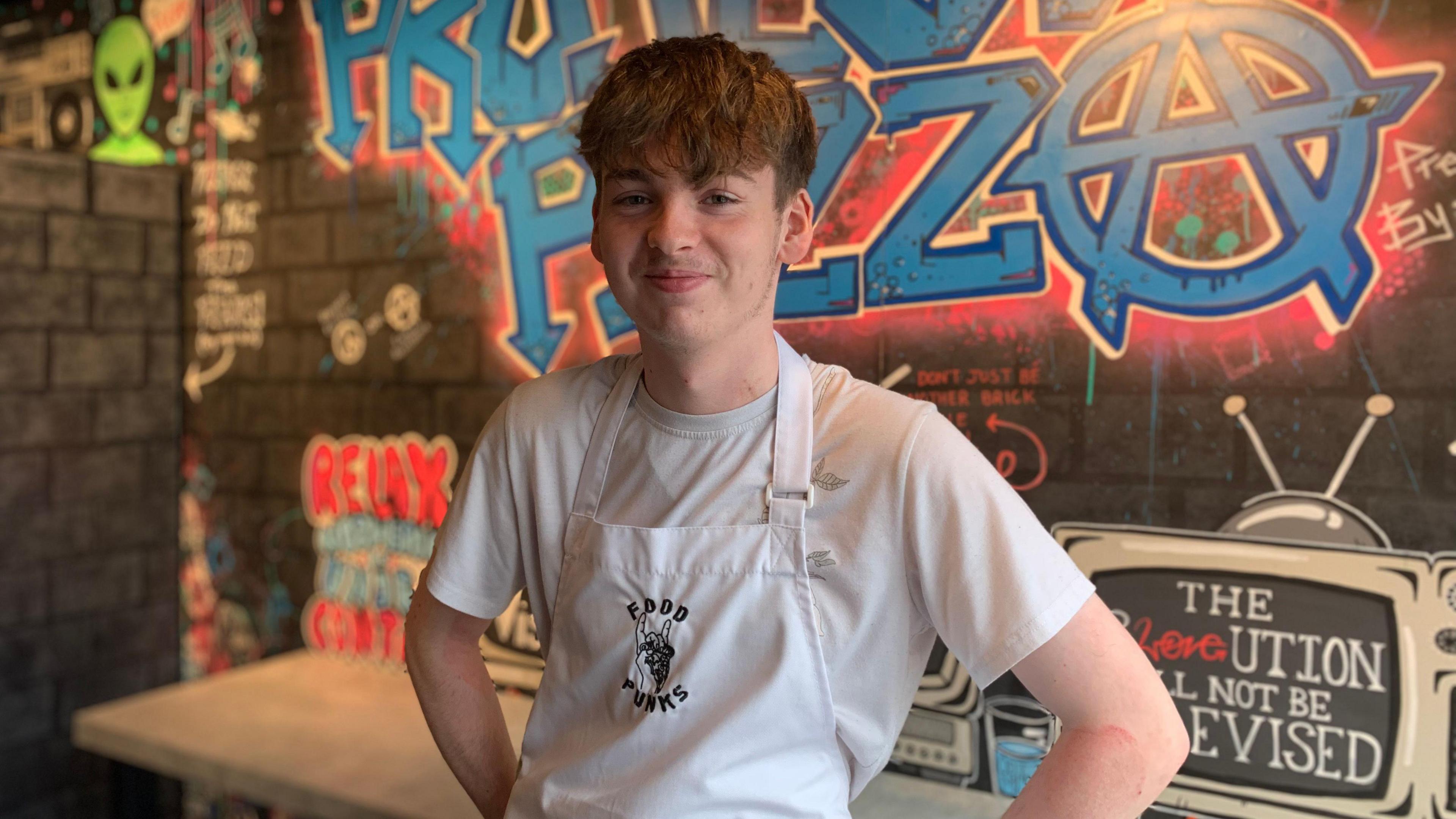 A young man in an apron and white t-shirt with dark hair stands smiling in front of a sign reading Project Pizza