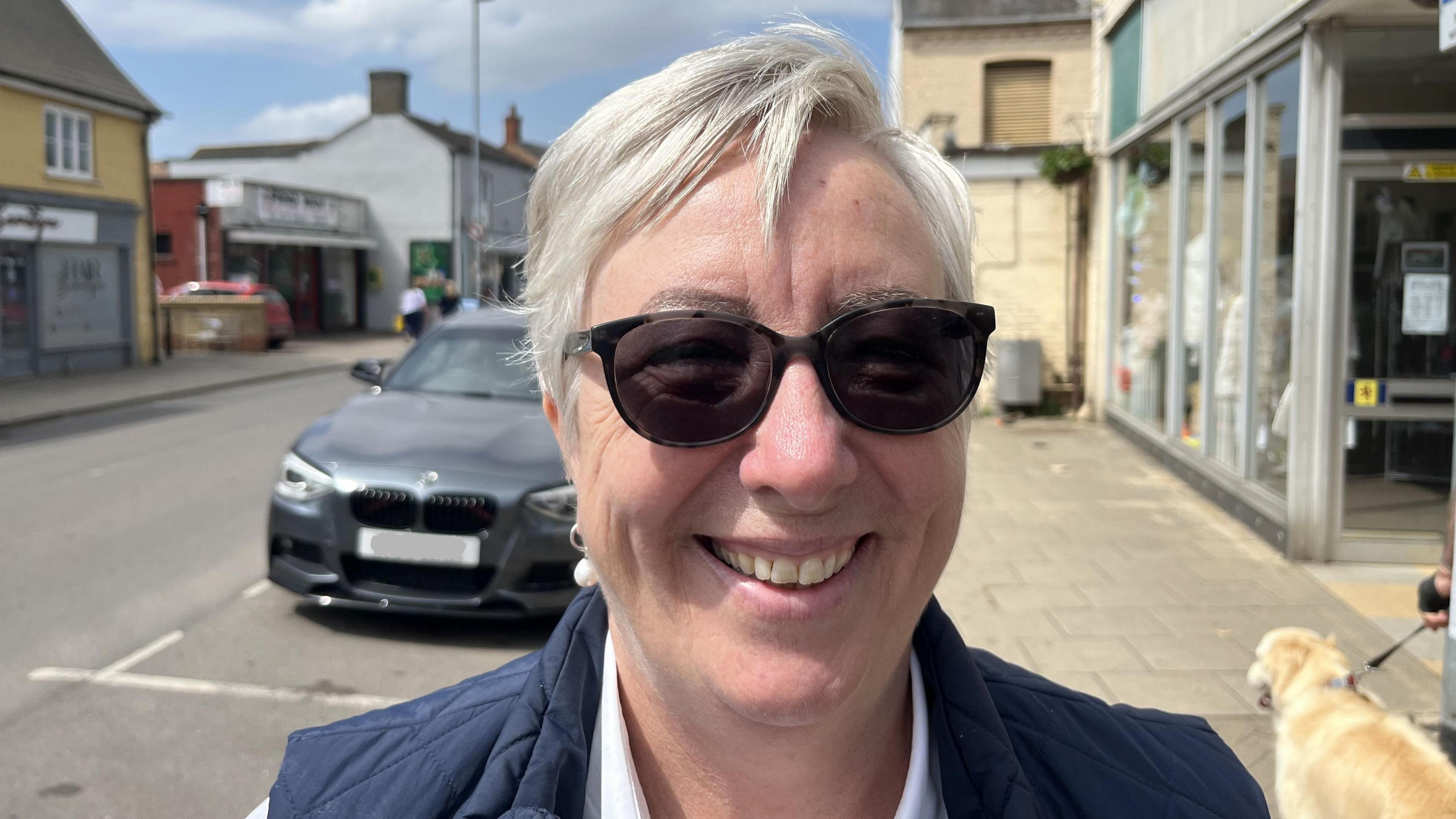 Jayne Brian-Platt smiling at the camera on the High Street with a dog and car in the background. She has short grey hair and is wearing tinted glasses and a navy blue bodywarmer.