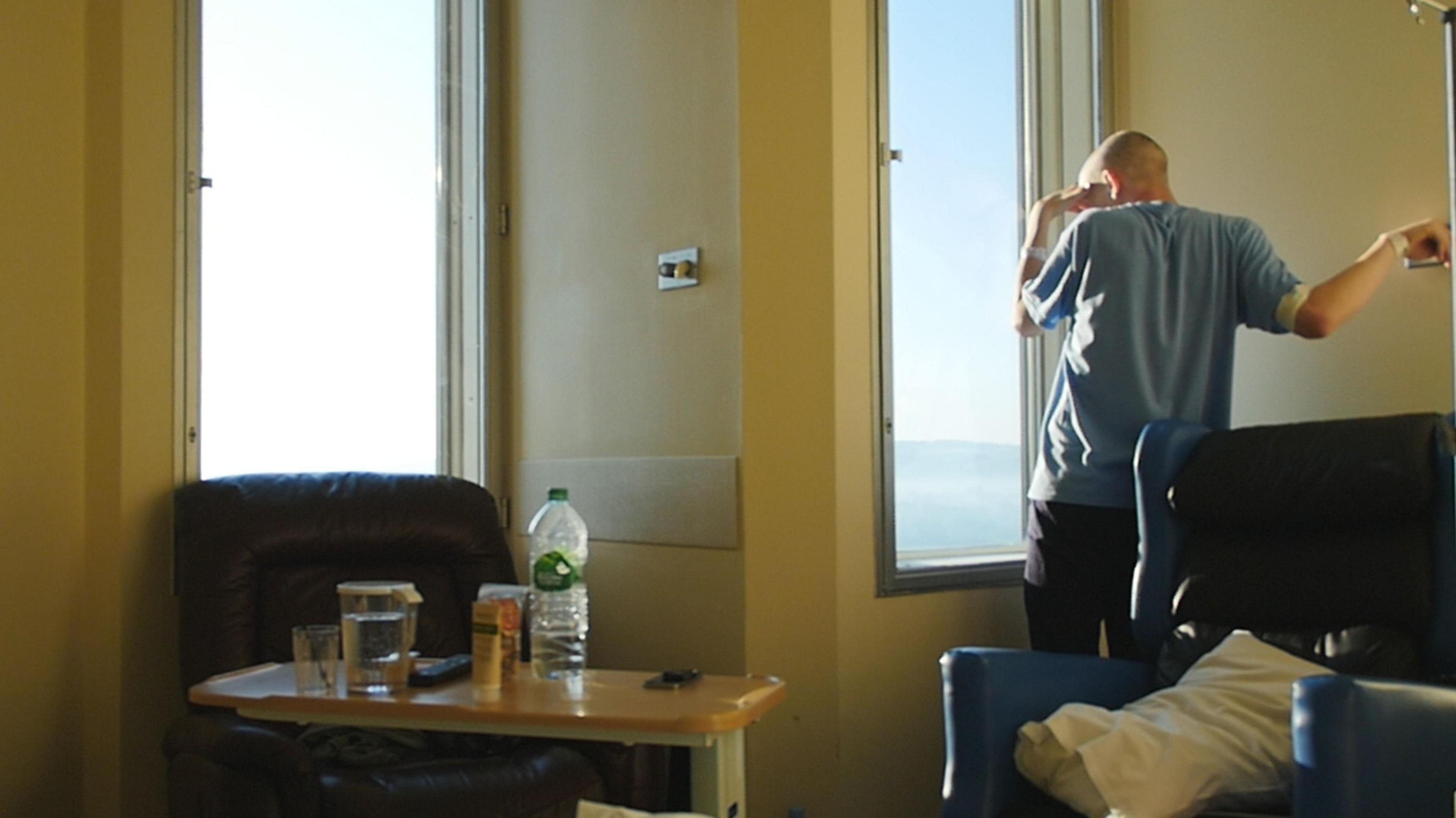 Éanna Mac Cana has his back turned to the camera as he stares out of the window of his hospital room. He is bald, and is wearing a light blue t-shirt and dark trousers. There are two armchairs in the room - one brown on the left and one dark blue on the right. There is a small table sitting in front of the left armchair with an empty bottle of water, a glass of water and a TV remote. Éanna has his arm up and is holding onto a metal pole and he brings his hand over his eyes.