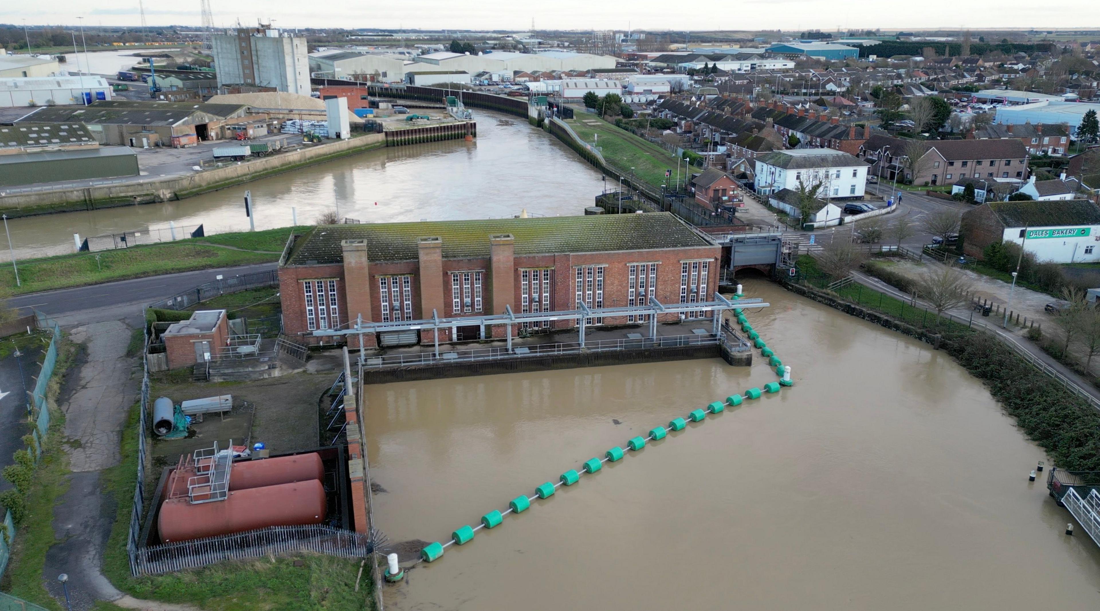 An aerial image of a waterway and pumping station in Boston, Lincolnshire. The red brick building stretches across the water and is surrounded by green floats. A number of houses and industrial buildings are visible in the distance.