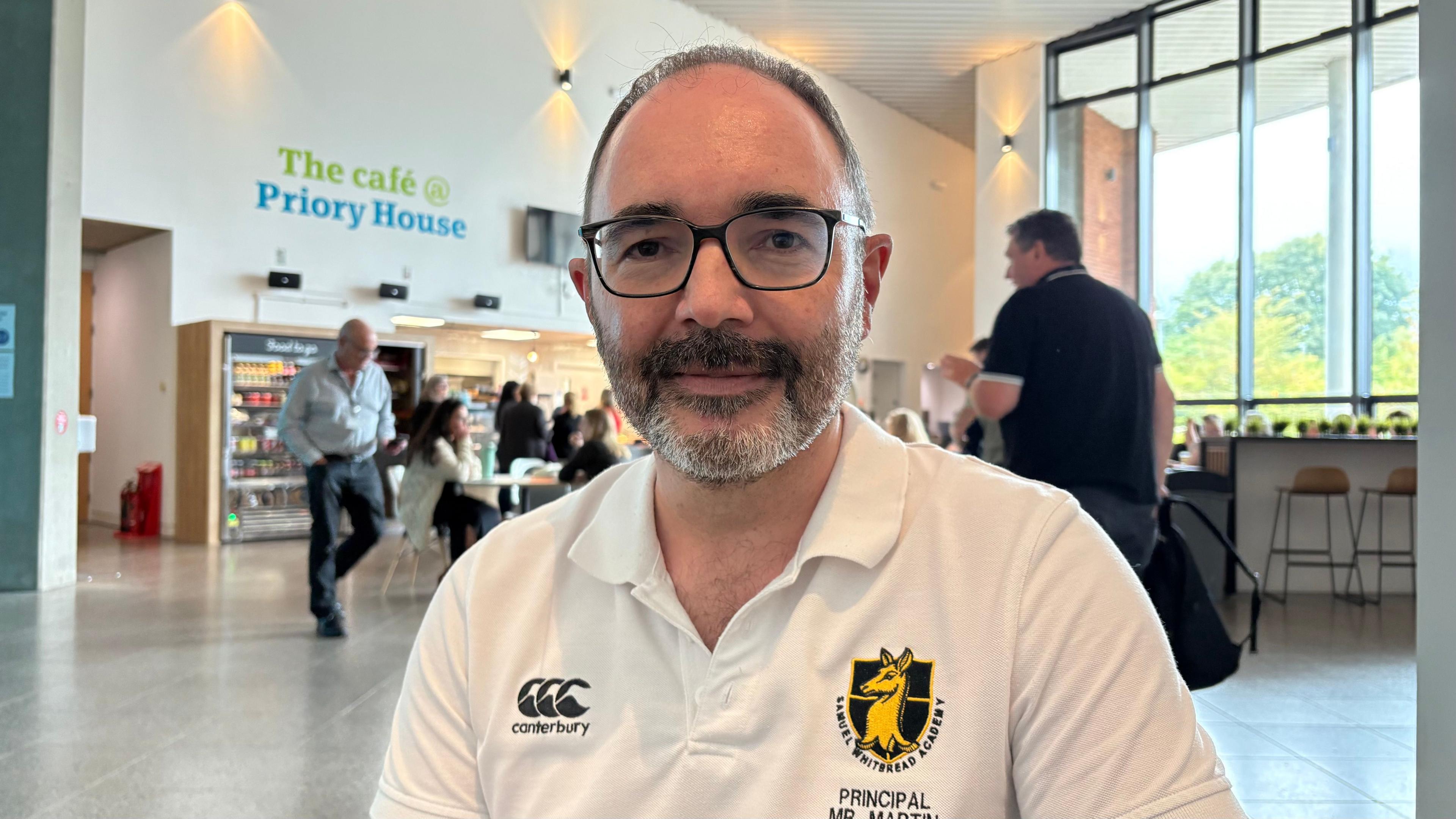 Nick Martin with short dark hair and beard wearing a white T-shirt with his name and title on the front, looking at the camera with a cafe in the background
