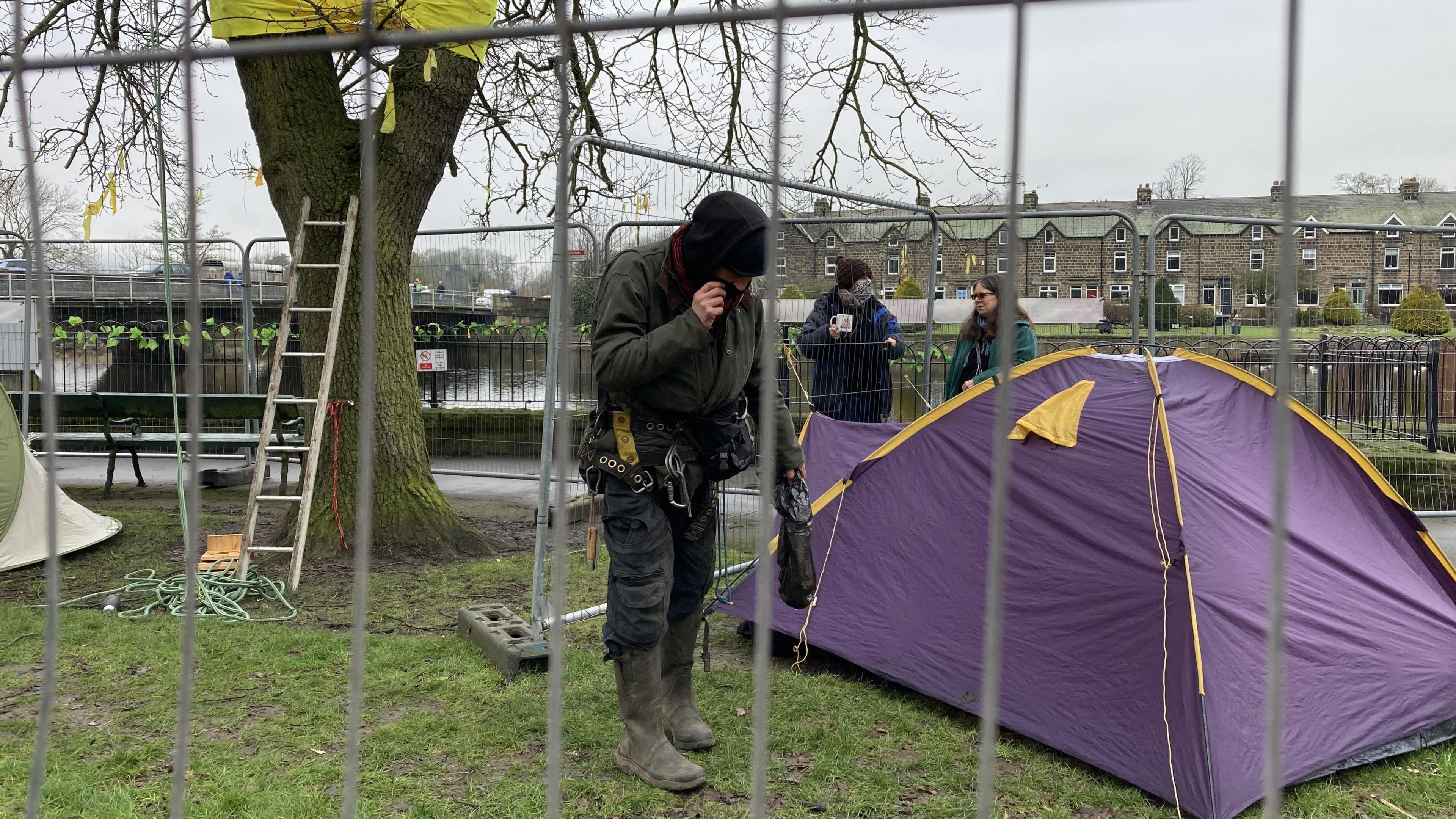 A purple tent sits behind a metal fence. A man dressed in a black hat, coat and outdoor clothes stands next to it.