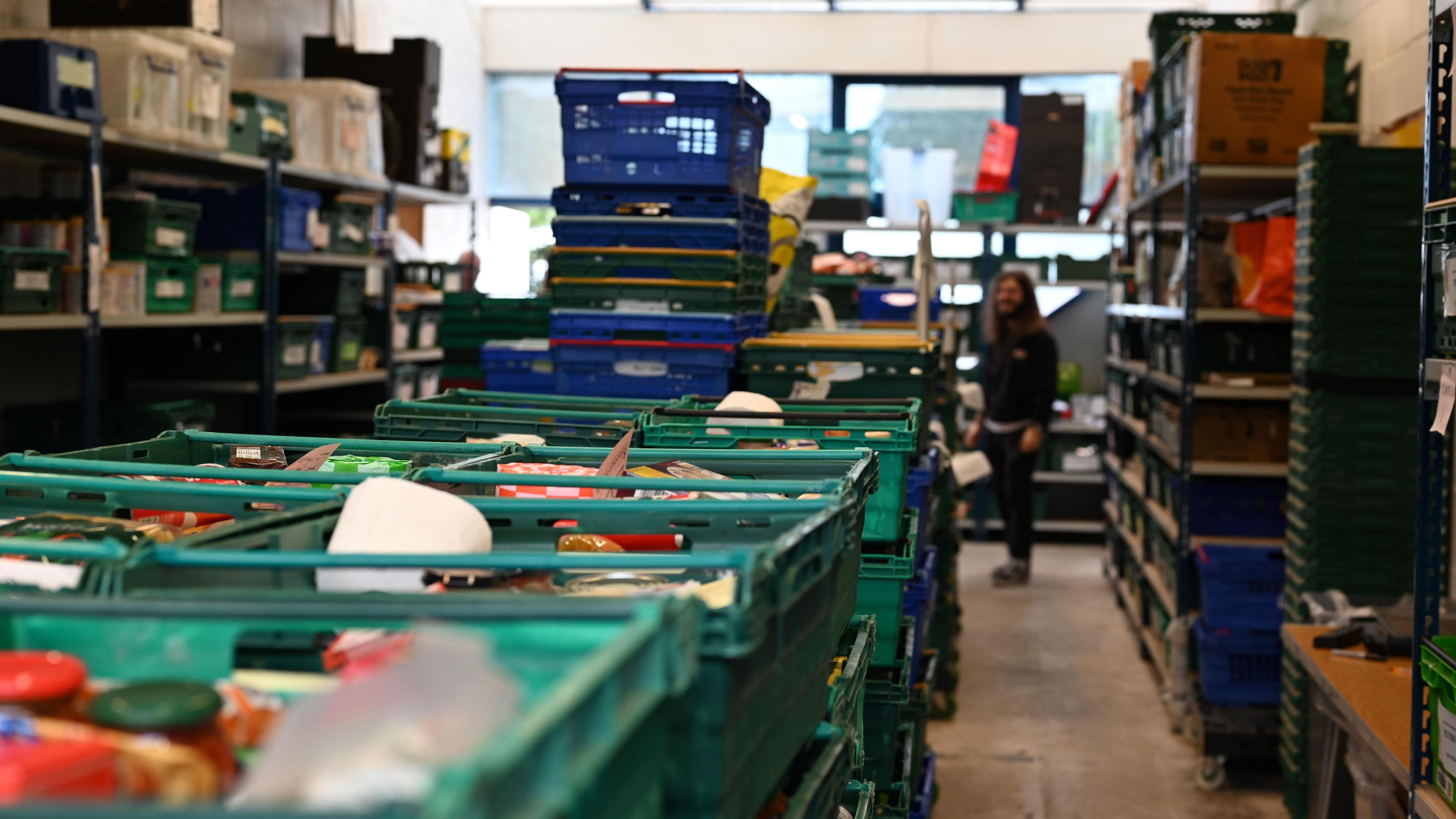 In the centre of a rectangle shaped room are stacks of green plastic trays. These are the food parcels given to foodbank users. At the sides of the room are shelves with more trays on. A person stands in the background holding a piece of paper while looking at the food parcels.