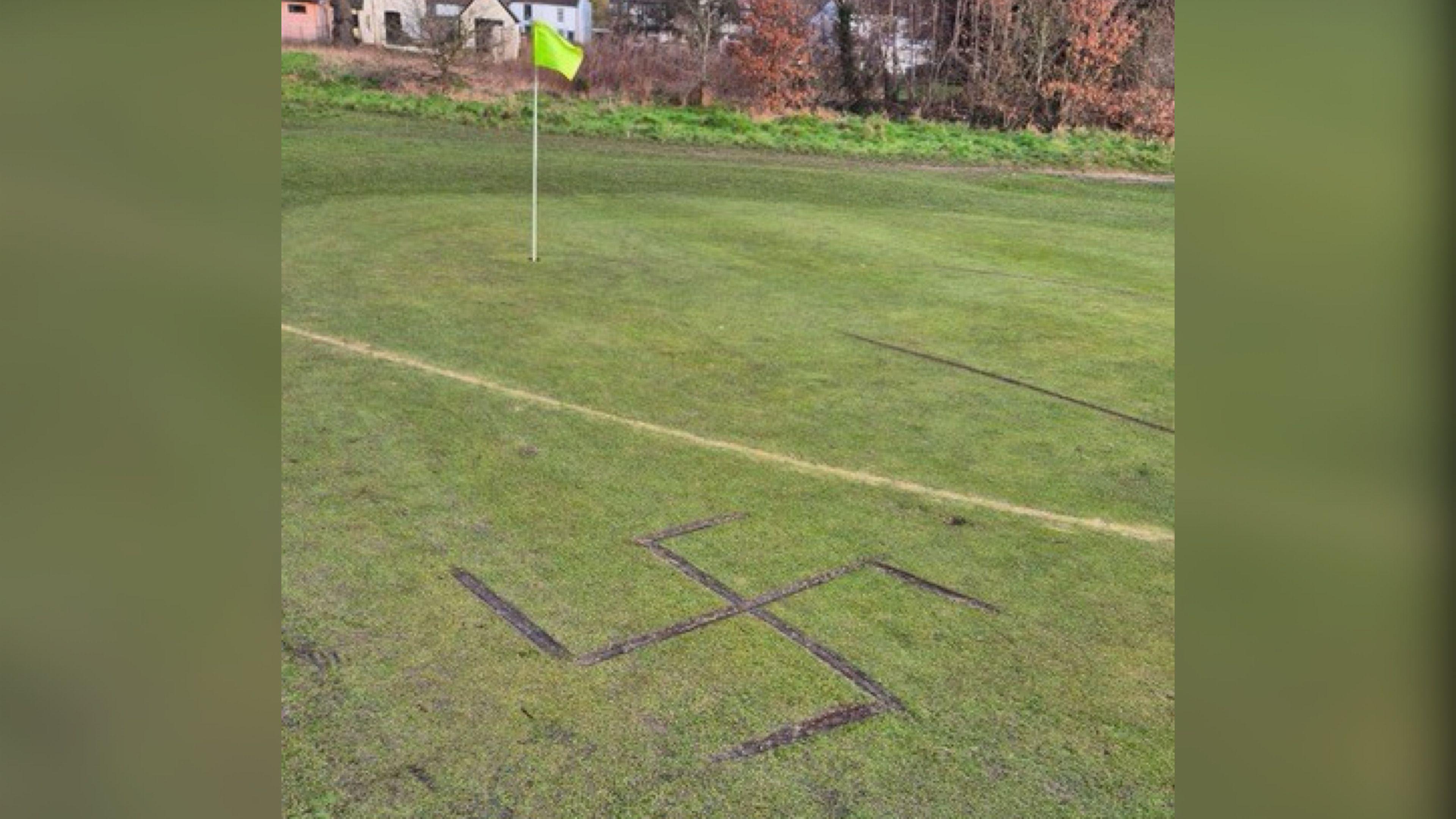 A golf green with a flag in the background which is hi-vis yellow. In the foreground there is a swastika which has been carved into the ground.