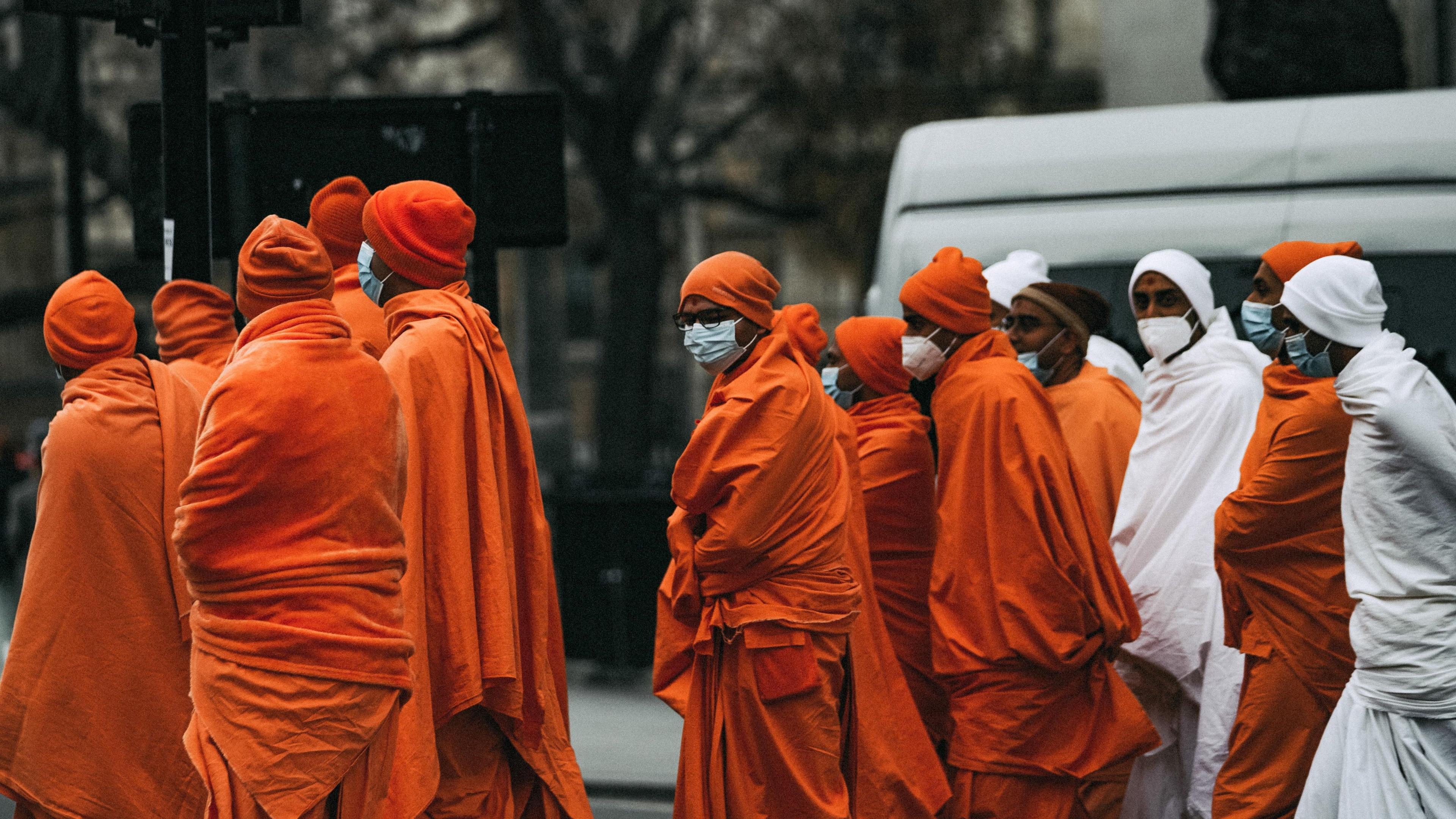 12 men in robes, mostly orange but some in white, crossing a road. They all have hats on in the same colour as their robes. They all wear masks. One in the centre stands out slightly as being slightly alone in the group.