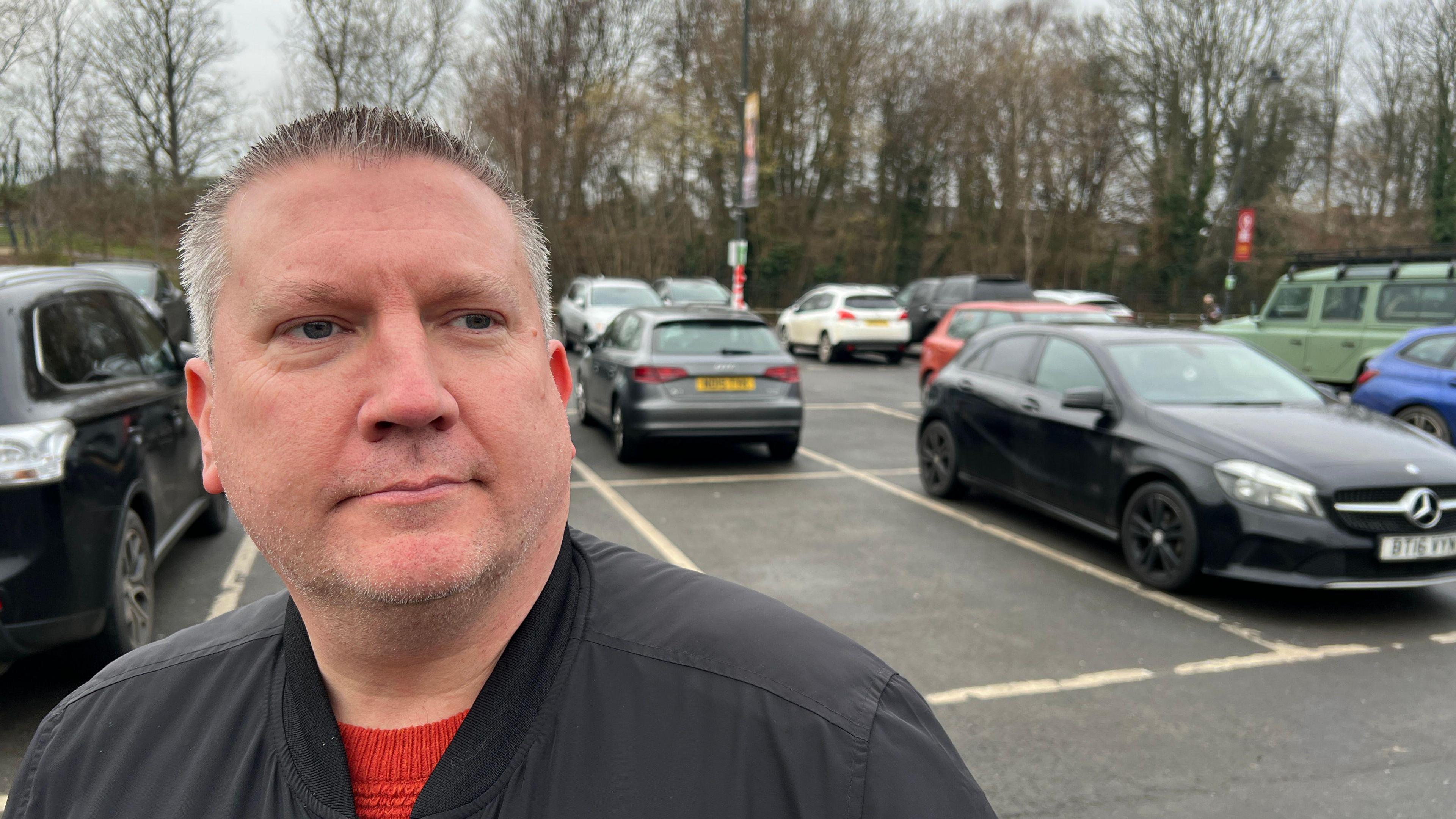 York's Chris Marsden is looking to the right of the camera. He has short, grey hair and is wearing a black jacket and red jumper. York's Marygate car park is in the background.