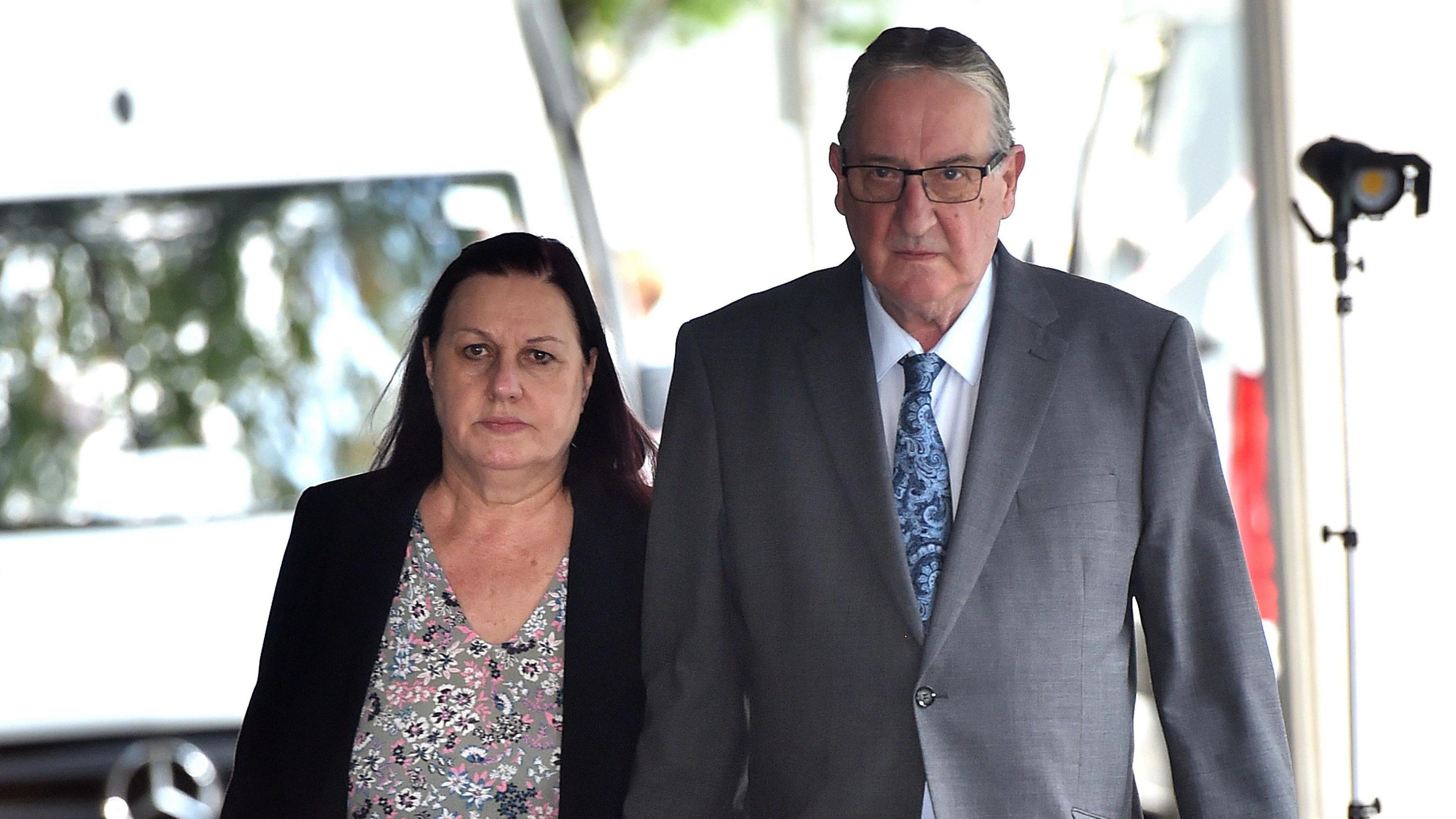 Susan Letby, with dyed red hair and wearing a black blazer over a flowery blouse, and John Letby, who has grey hair, glasses and wears a grey suit with a blue tie, walk towards the camera with a serious expression 