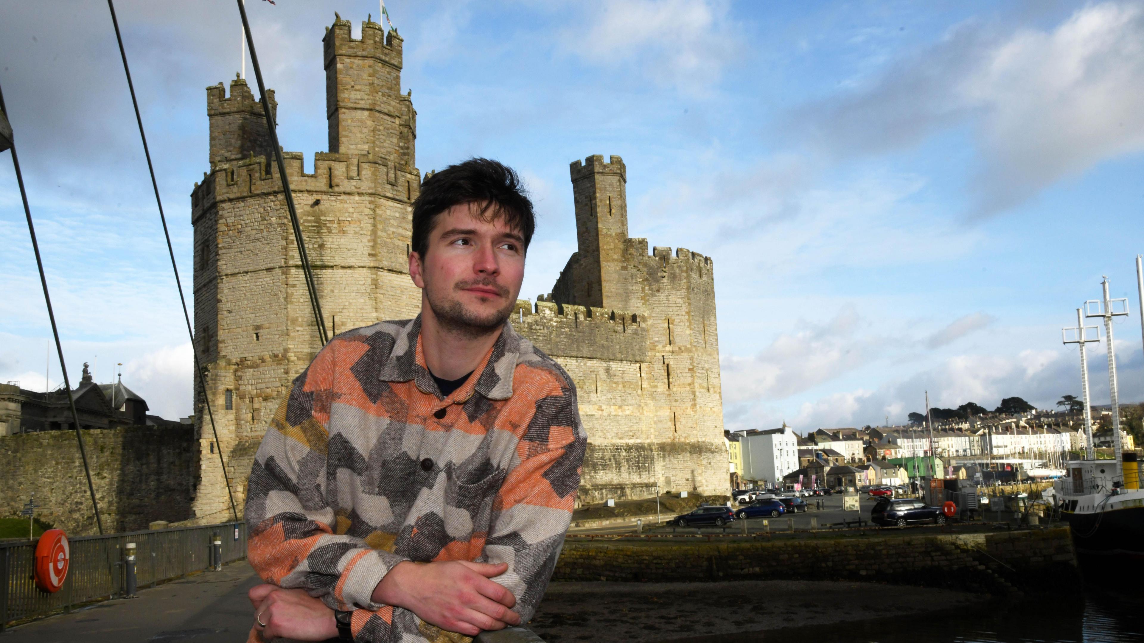 Iestyn Tyne yn sefyll ar Bont yr Aber gyda Chastell Caernarfon yn y cefndir