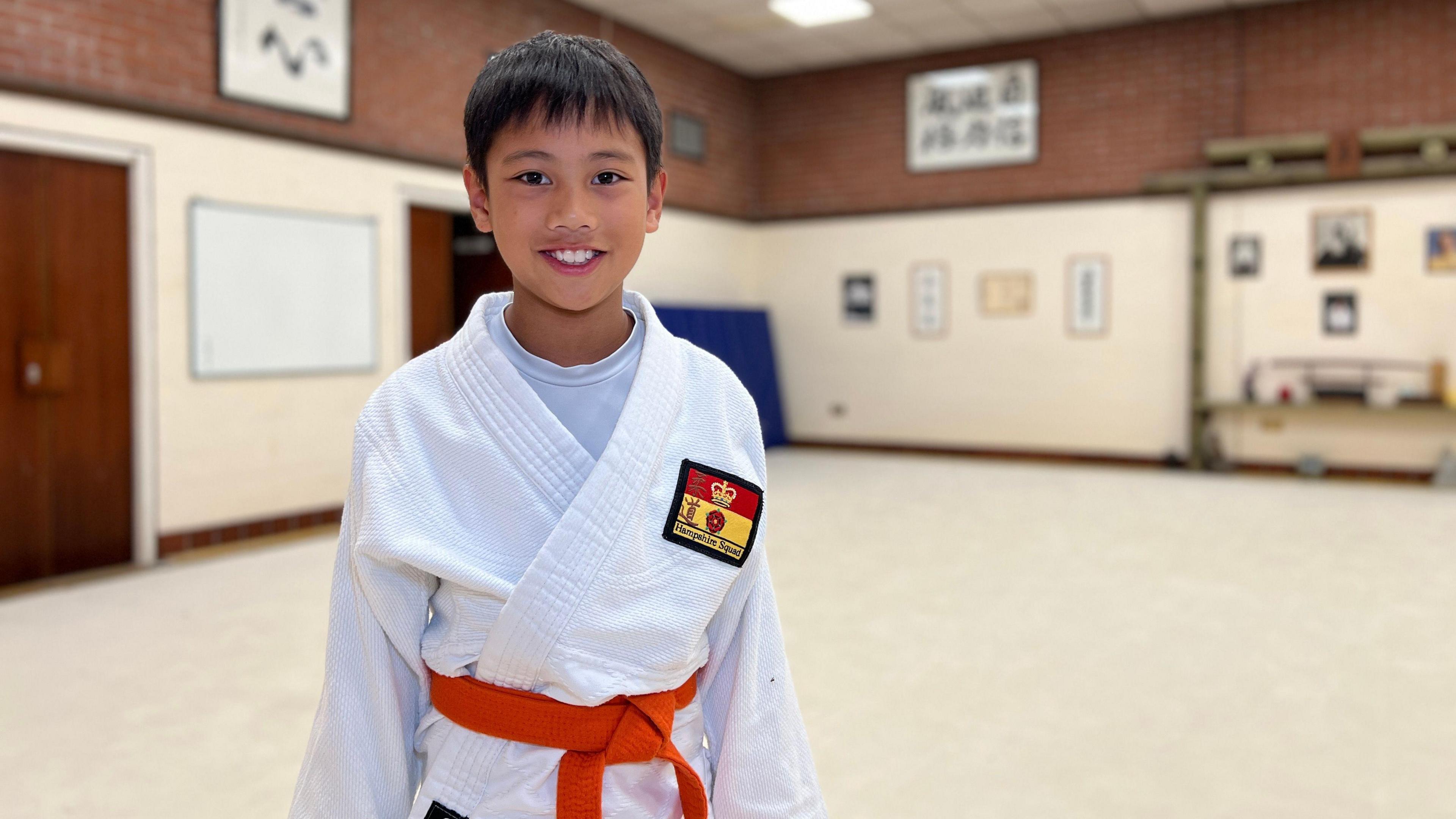 Lucas smiles at the camera, he has brown hair and brown eyes. His hair covers most of his forehead. He's wearing his judo gi and has a Jersey Judo badge sown on to the righthand side of his chest. Lucas is wearing an orange belt.