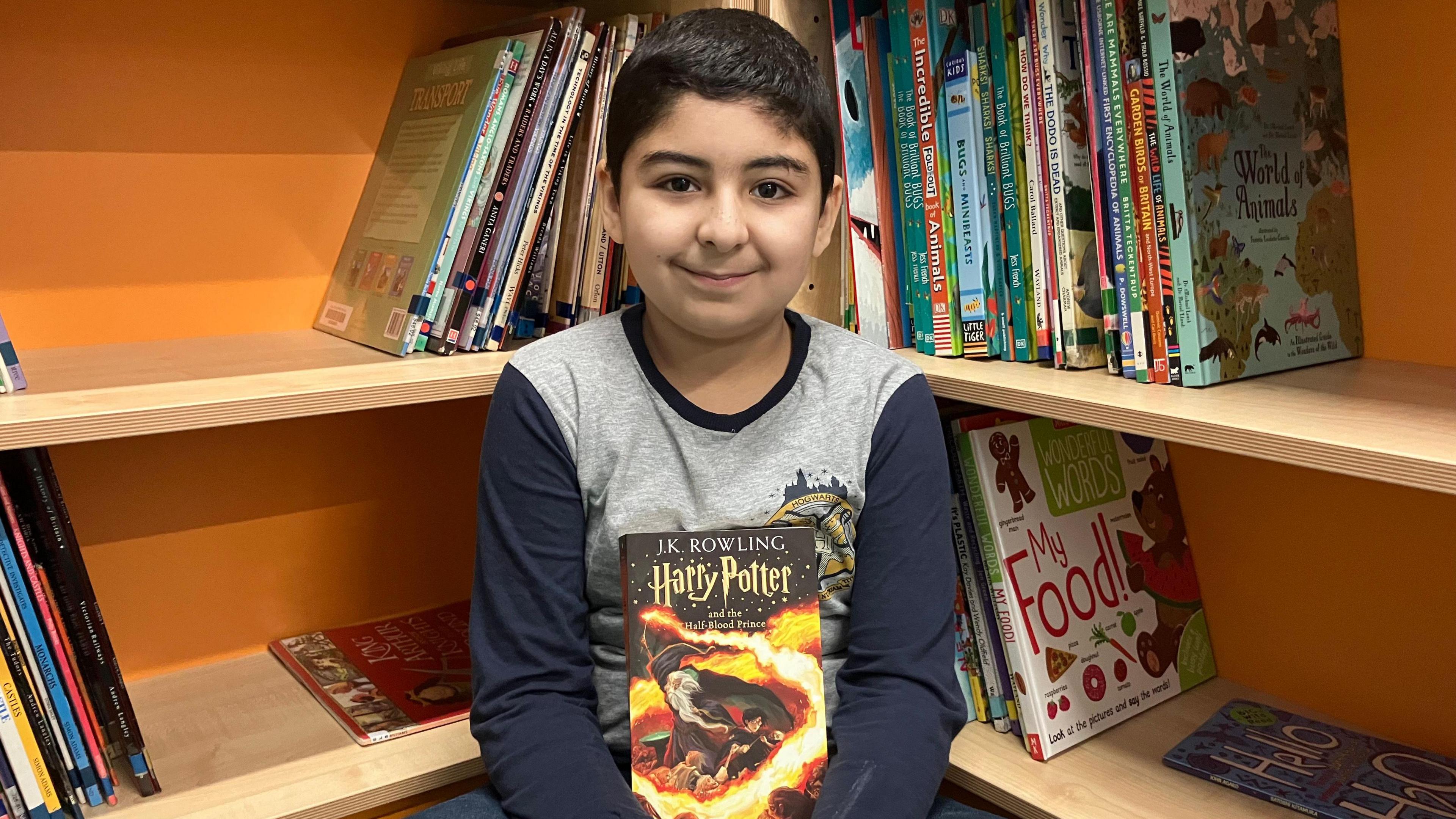 Mustafa, who is wearing a blue and grey shirt, sat in front of bookcases, which are at a right angle, with a copy of a Harry Potter book. 