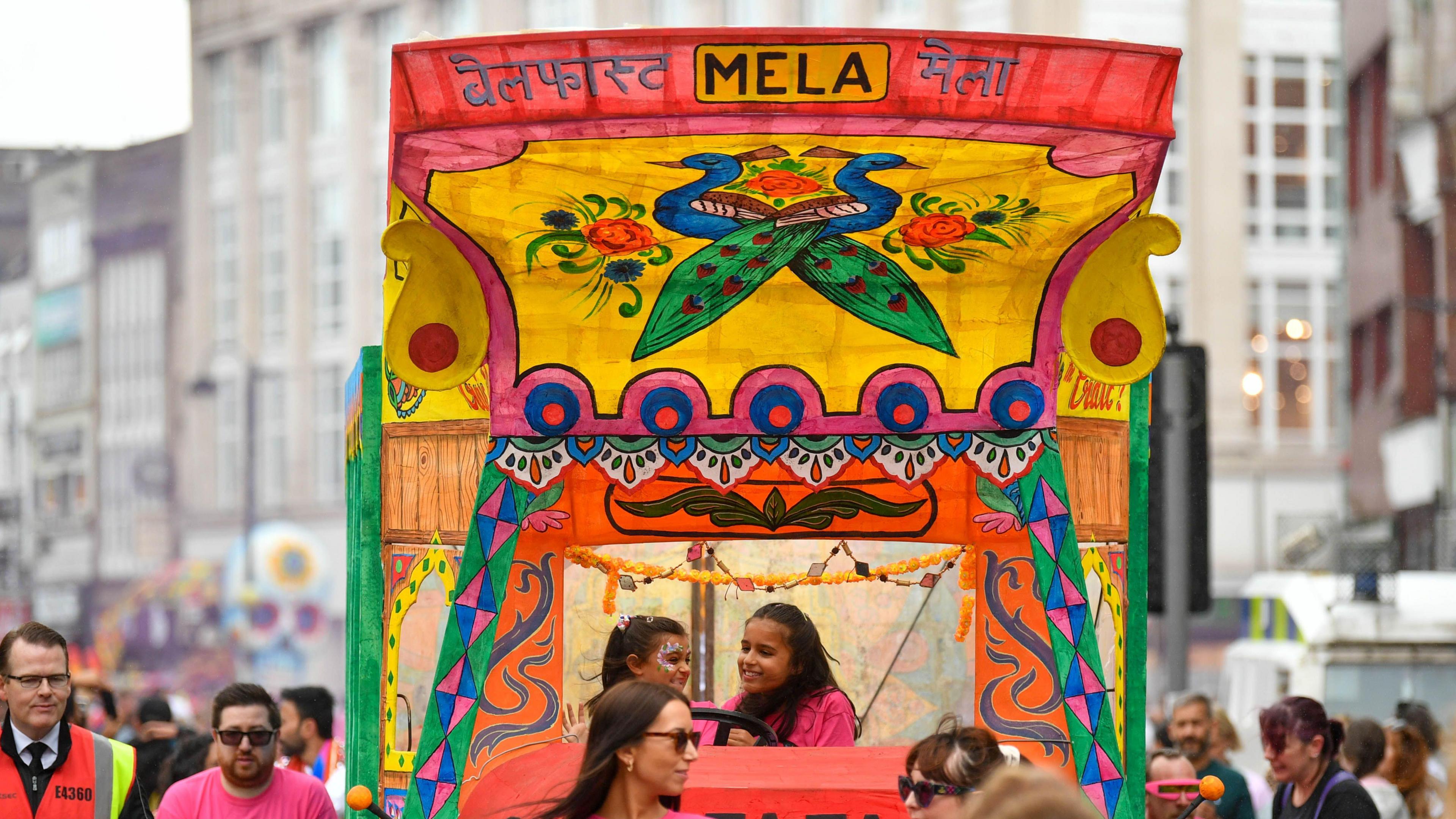 Two young girls on a Mela float, which has loads of colour and has peacocks on the front of the cover 