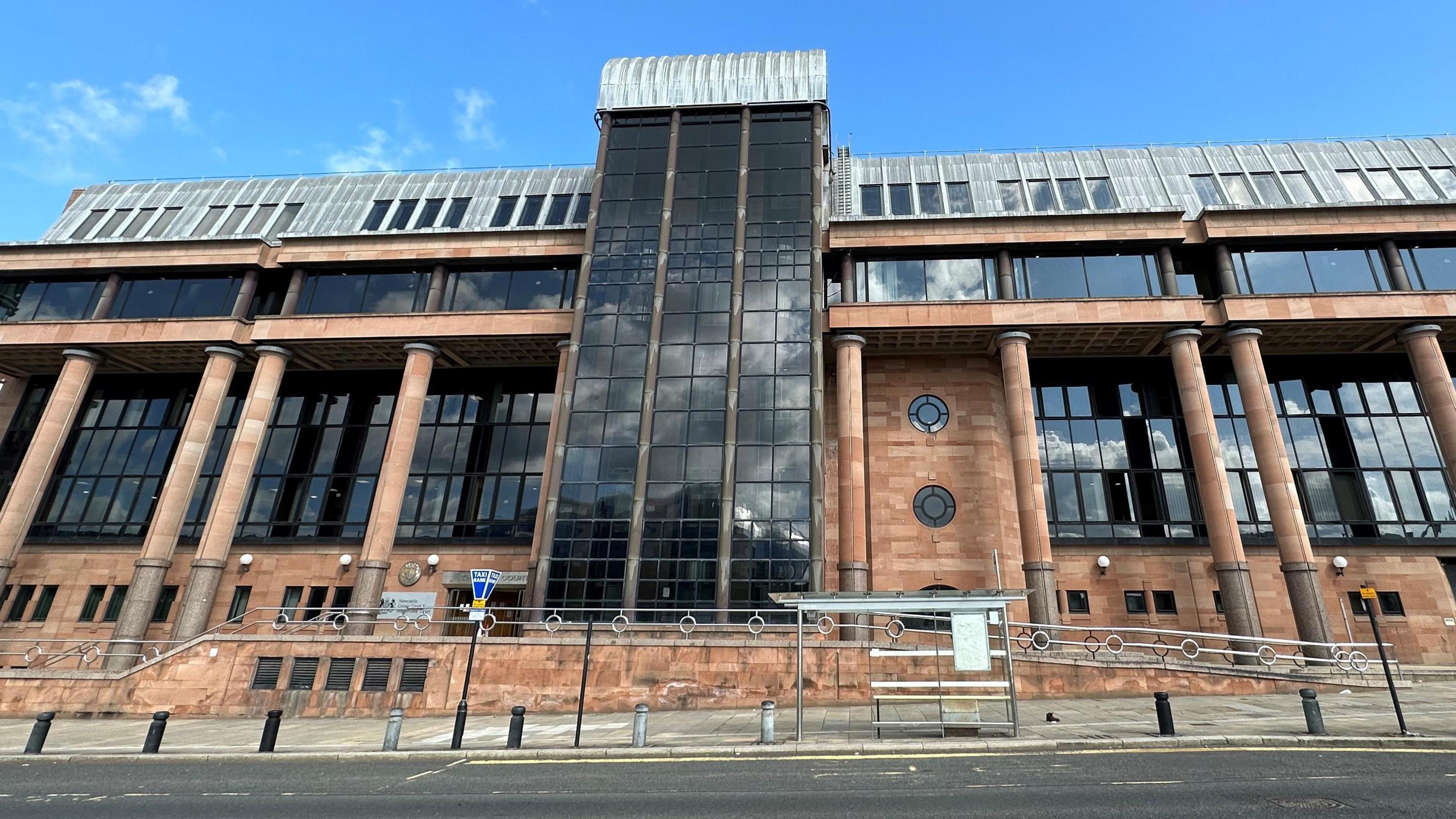 Newcastle Crown Court. It is a large imposing building with red brick walls and columns and large dark windows.