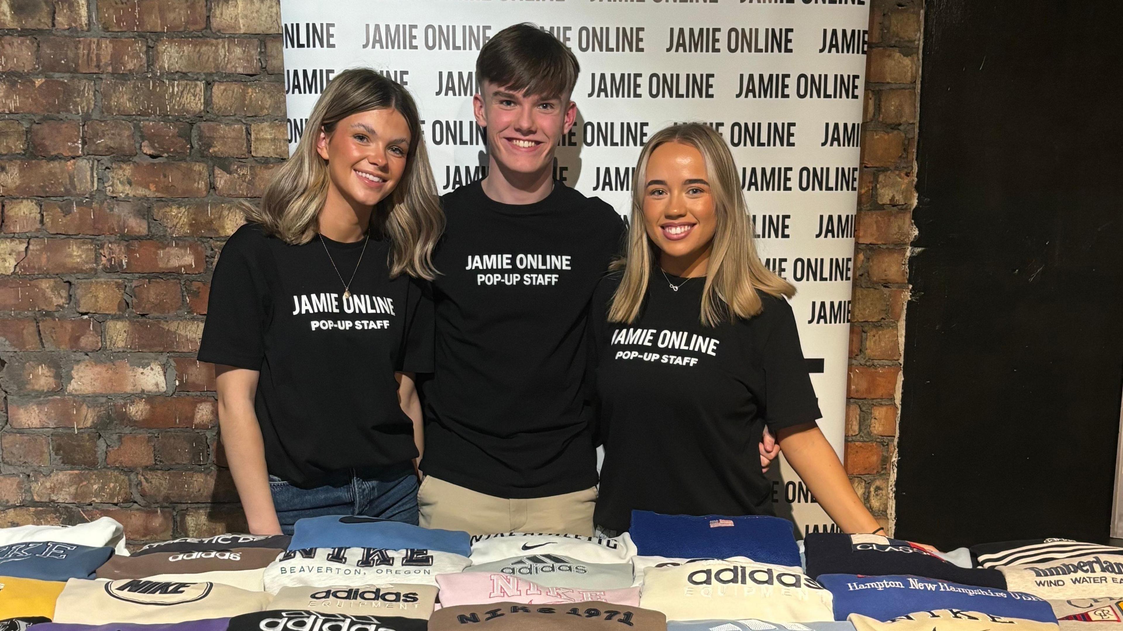 Two females and one male standing in front of a Jamie Online banner, the banner is white and in front of a red brick wall. In front of the three people is a table covered completely in retro sweatshirts there is 28 in total. To the right of the image is a poster of influencer Molly Mae wearing a sweatshirt,  