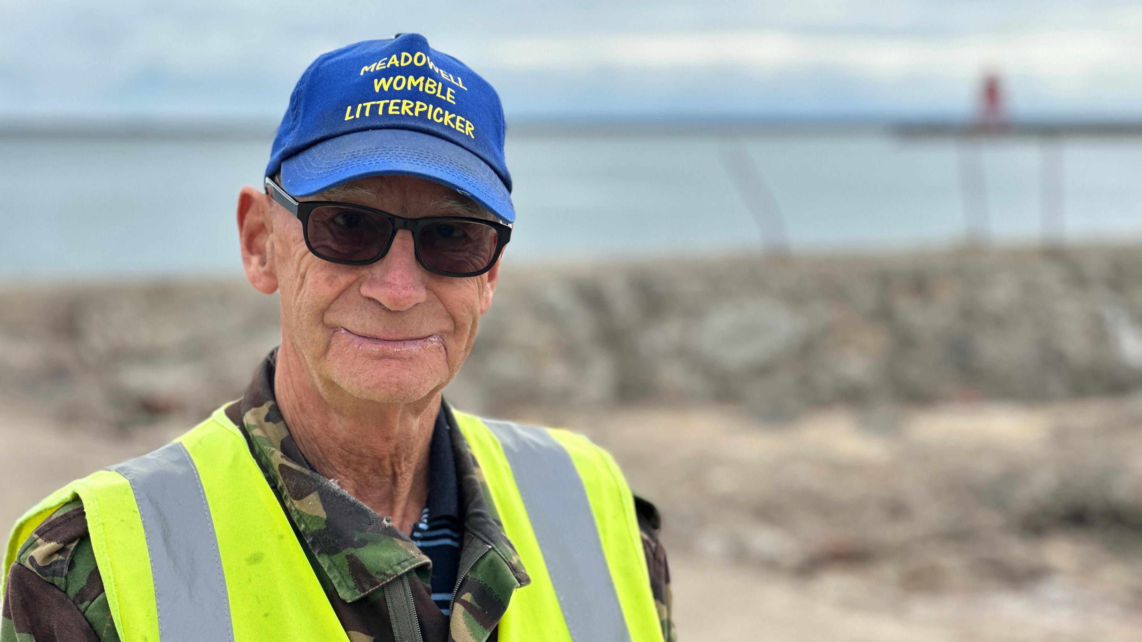 Dave Shaw is standing facing away from the beach and water, which are out of focus behind him. He is wearing a blue baseball hat with yellow lettering saying he's a "Womble litter picker" and is wearing a high-vis jacket over a camouflage jacket. He is wearing sunglasses and is half smiling.