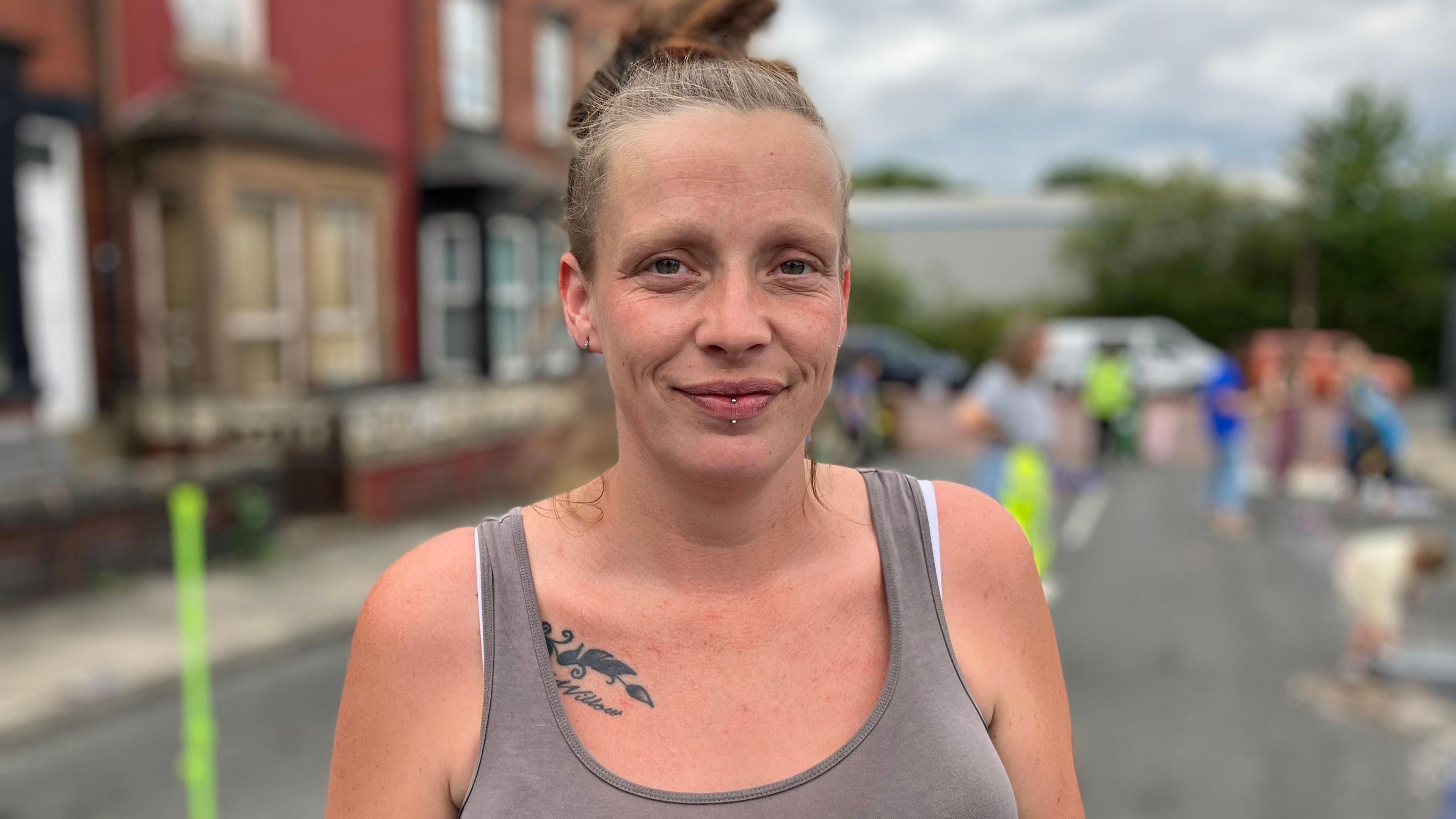 A achromatic pistillate wearing a grey vest apical is smiling astatine the camera. She is wearing her blonde hairsbreadth up successful a bun. Behind her children are playing successful a residential thoroughfare that has been blocked disconnected to traffic.