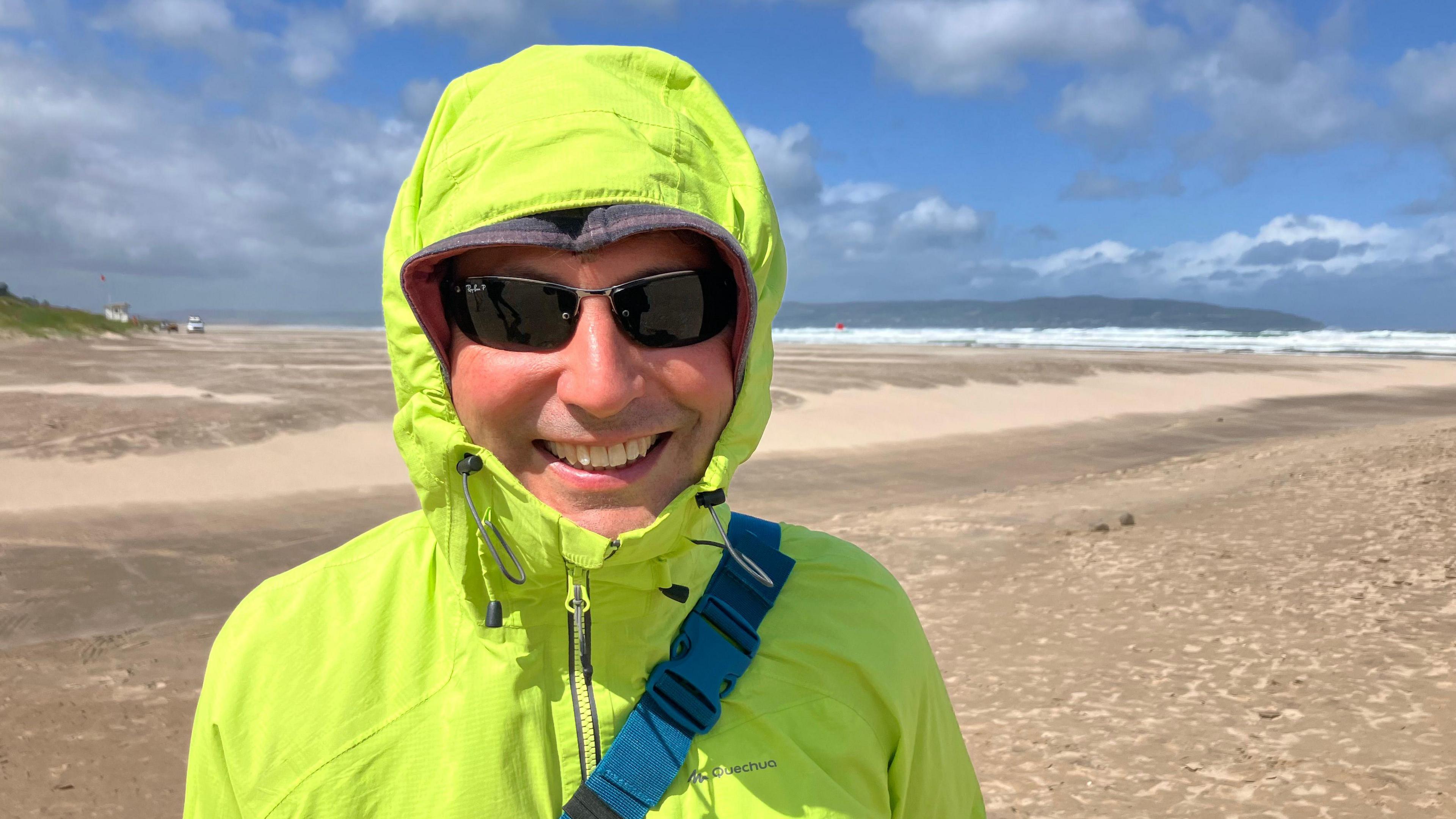 Martin. He is wearing a yellow fluorescent rain coat and sunglasses. The beach is in the background.