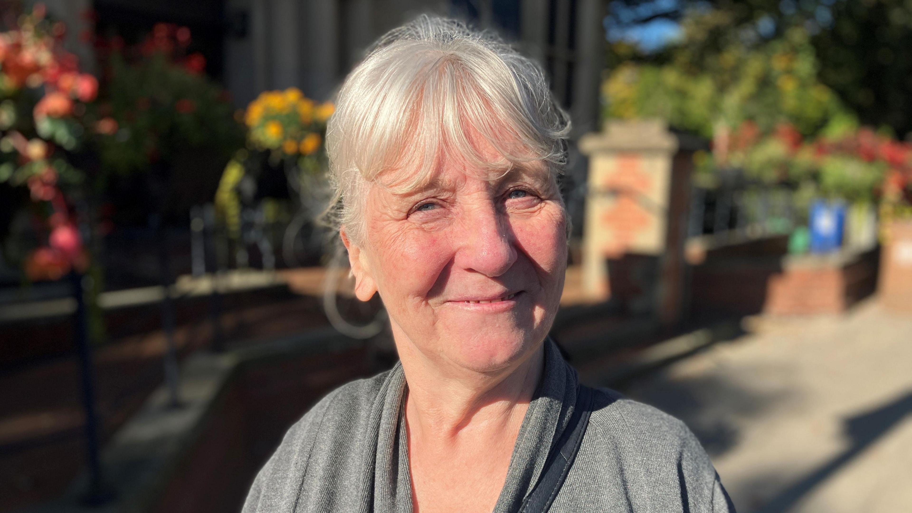 A photo of a middle-aged woman standing outside in the sun smiling. She is wearing a grey jumper. 