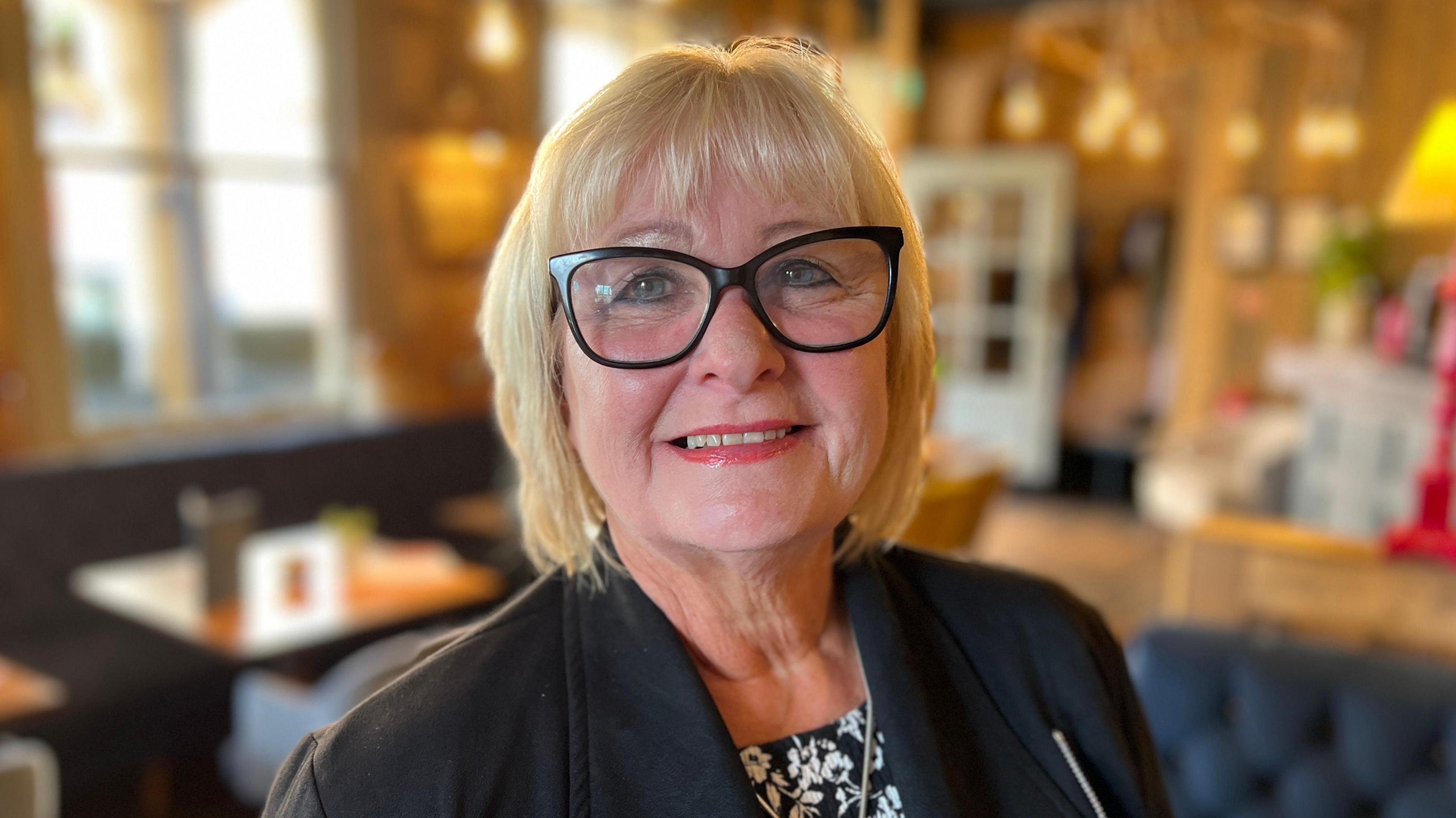 A middle-aged woman with glasses looks at the camera with a hotel interior blurred in the background.