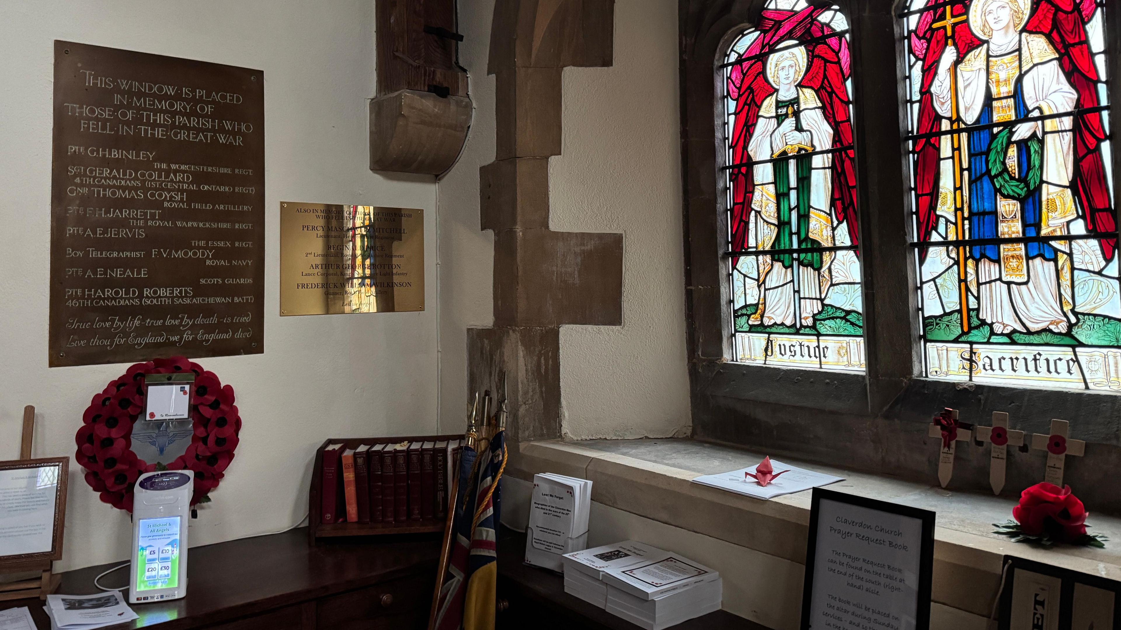 A new brass plaque is on the wall next to a darker memorial dating back to 1921. A poppy wreath has been put up after the Remembrance Day service. The memorials are in a corner next to two stained glass windows.