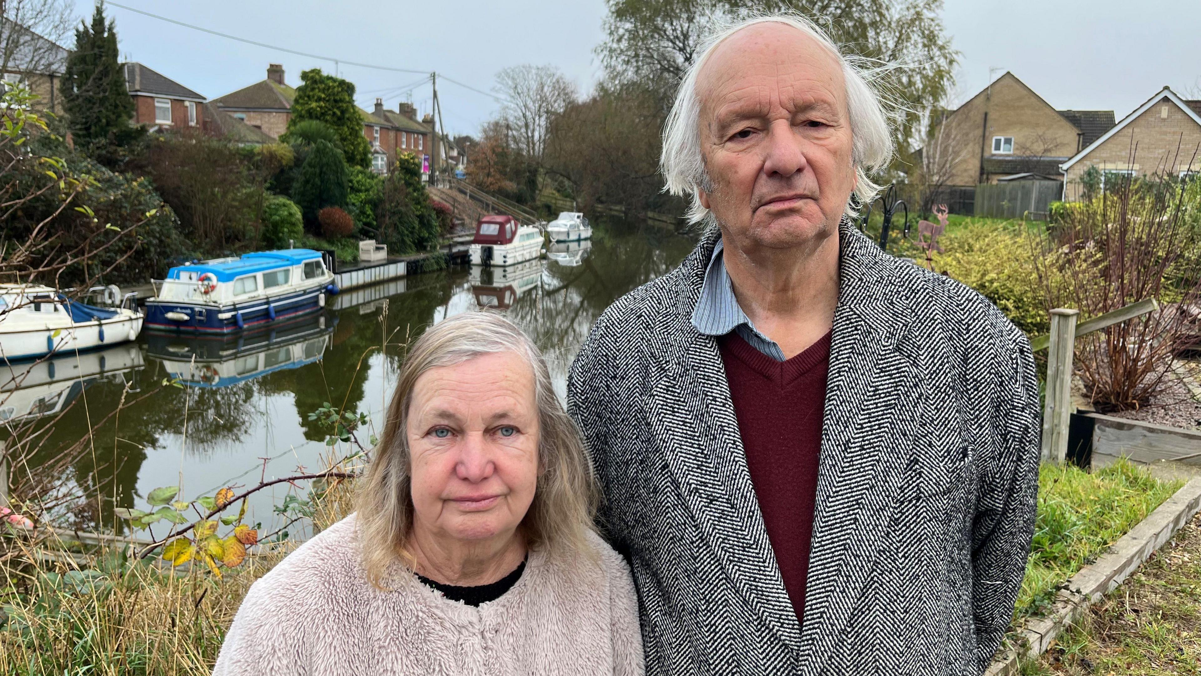 Tony Gosling, 75, on the right has grey hair and is wearing a herringbone black and white overcoat, with a purple jumper over a blue shirt, his wife, Patsy, 71, has grey and sandy coloured shoulder length hair parted to the left with a beige woollen fleece over a dark shirt. Behind them is a river with 4 cruiser boats moored on the far side, residential housing can be seen too.