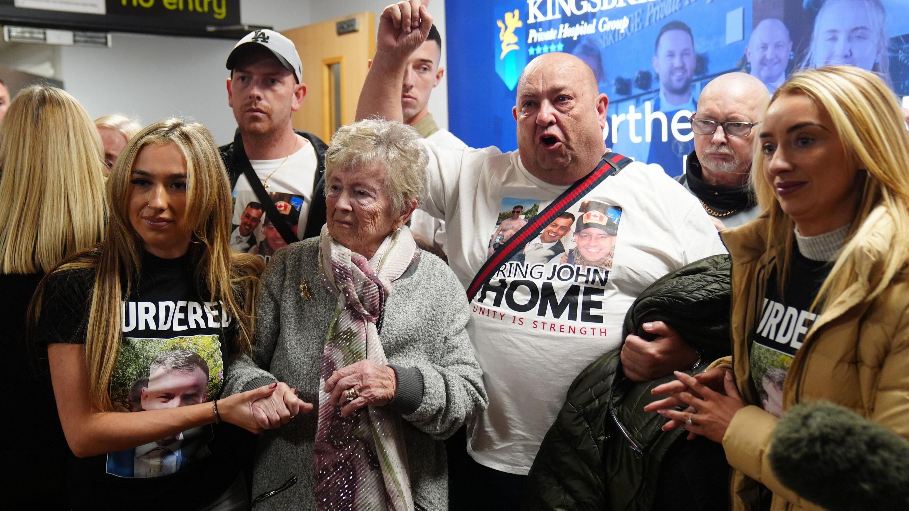 A man shouting and holding his finger in the air. He is surrounded by people, some of them wearing t-shirts with a picture of John George and the word "Murdered" printed on them