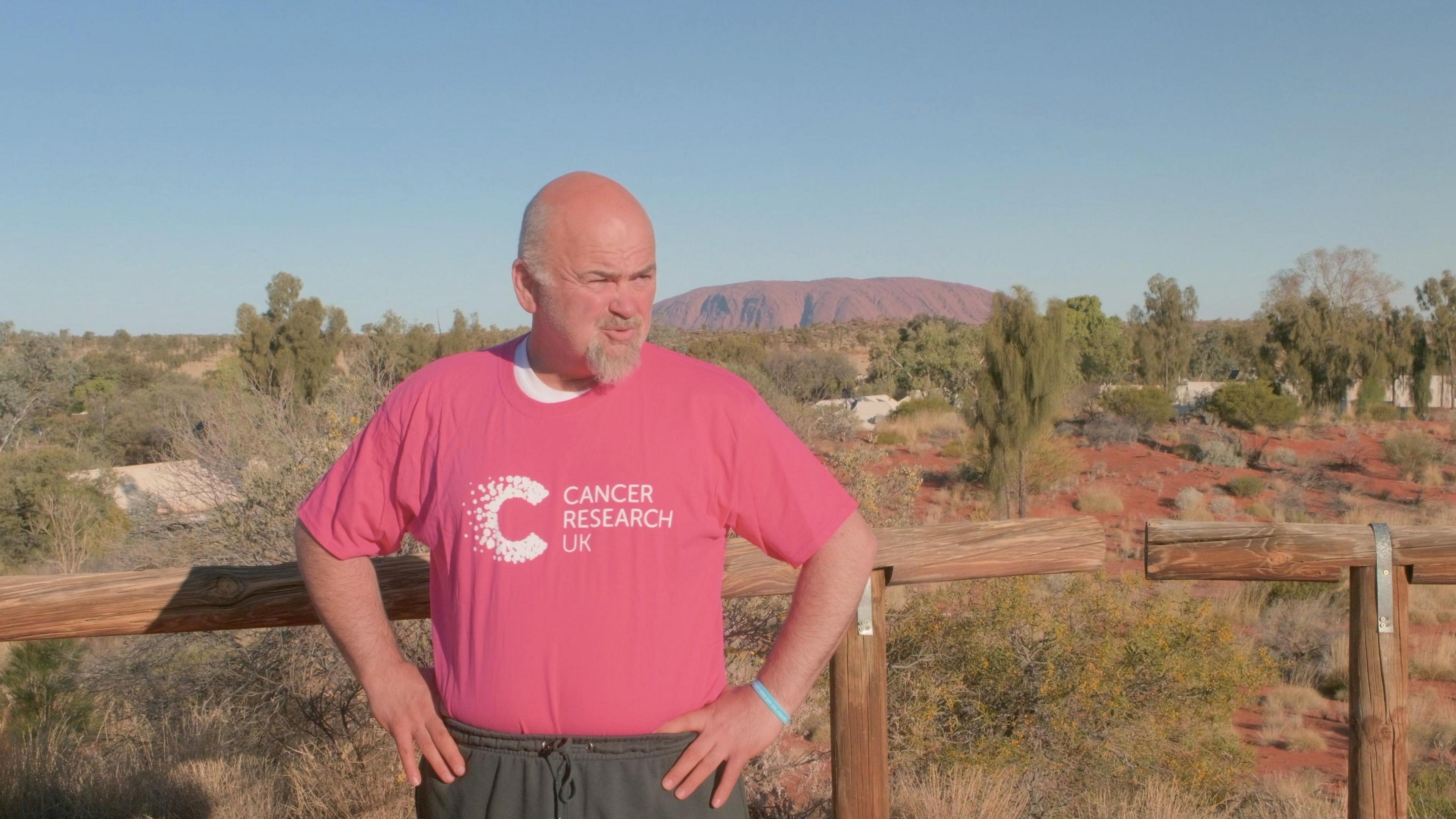 A bald man wearing a pink Cancer Research UK shirt and grey jogging bottoms stands in front of Uluru (Ayers Rock) in Australia. The large monolith is visible in the distance with several trees and patches of sand in front of it. The man has his hands on his hips and stands in front of a wooden fence.