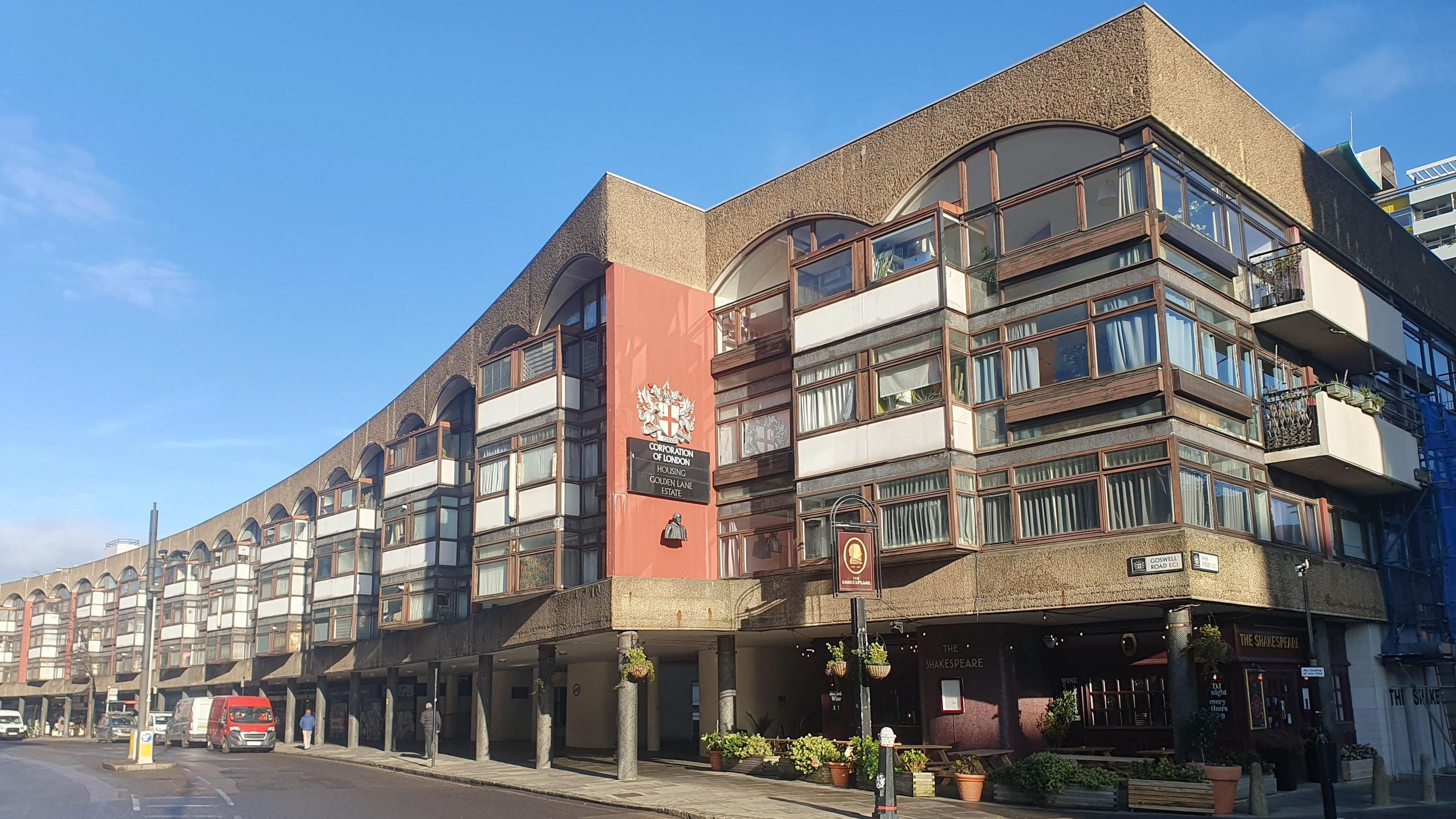 A large, modern building with a curved facade. The structure is multi-story, featuring wide windows and balconies on each level. Its exterior combines brick and concrete, with some areas painted red. Above an entrance, signage reads “Corporation of London" below the City of London Corporation crest. The sky is clear and blue.