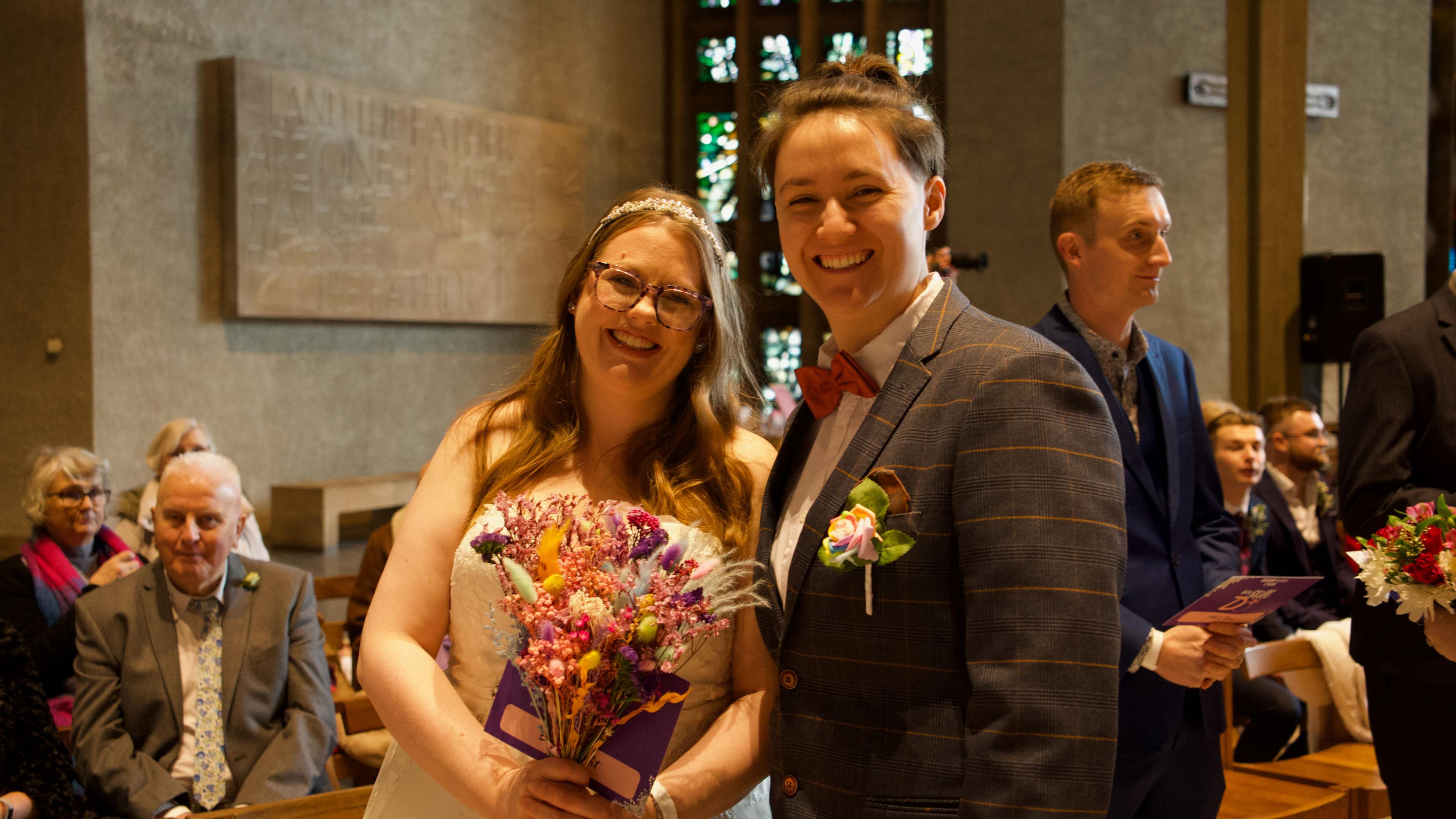 A woman in a white wedding dress and a bunch of flowers with her partner in a brown, check suit and red bow tie