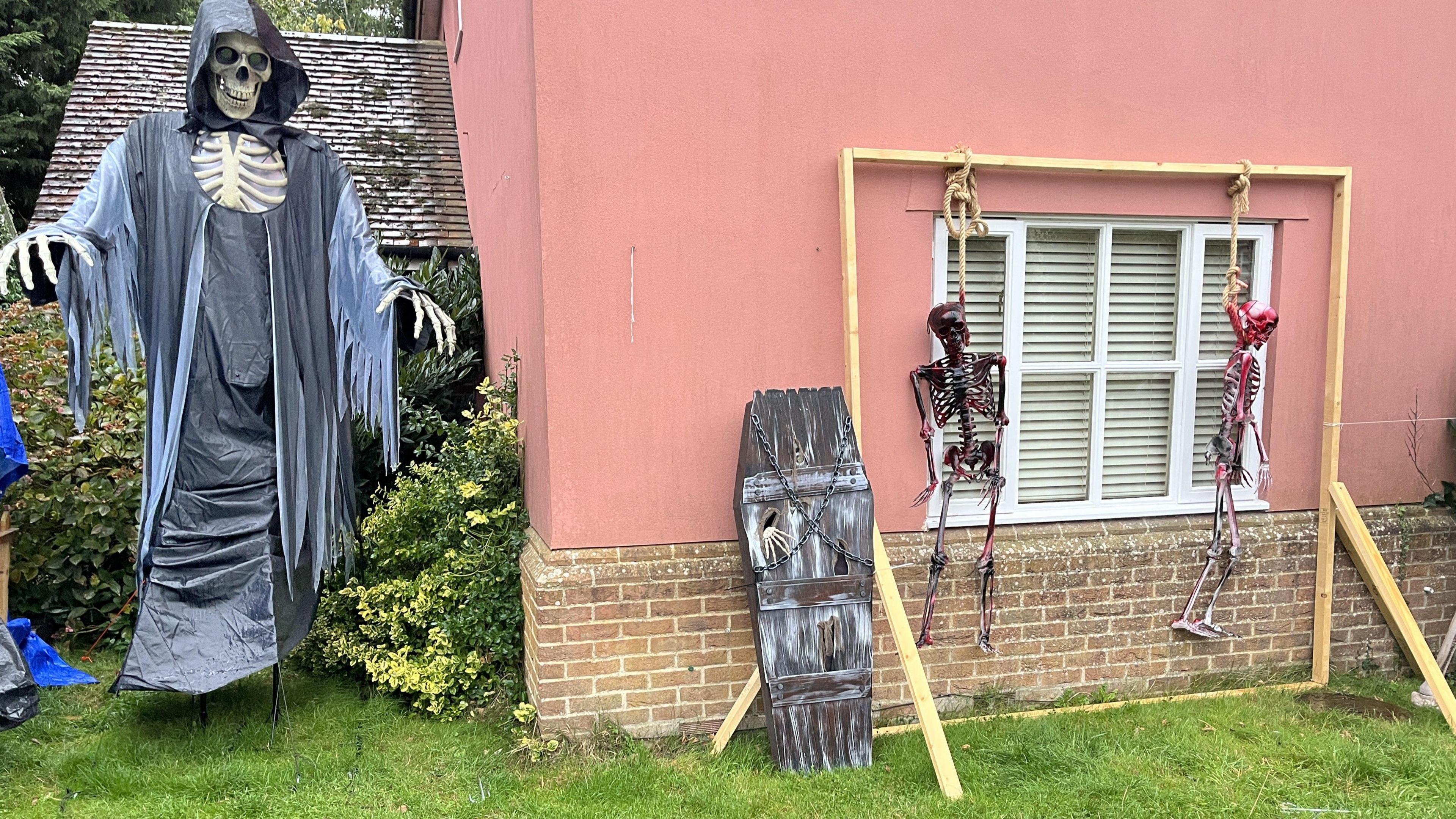 A skeleton wearing a grim reaper black cape stands to the left of the house. A coffin with a skeleton hand poking out the front rests against the front of the house. Two small red skeletons hang from a wooden frame in nooses. 