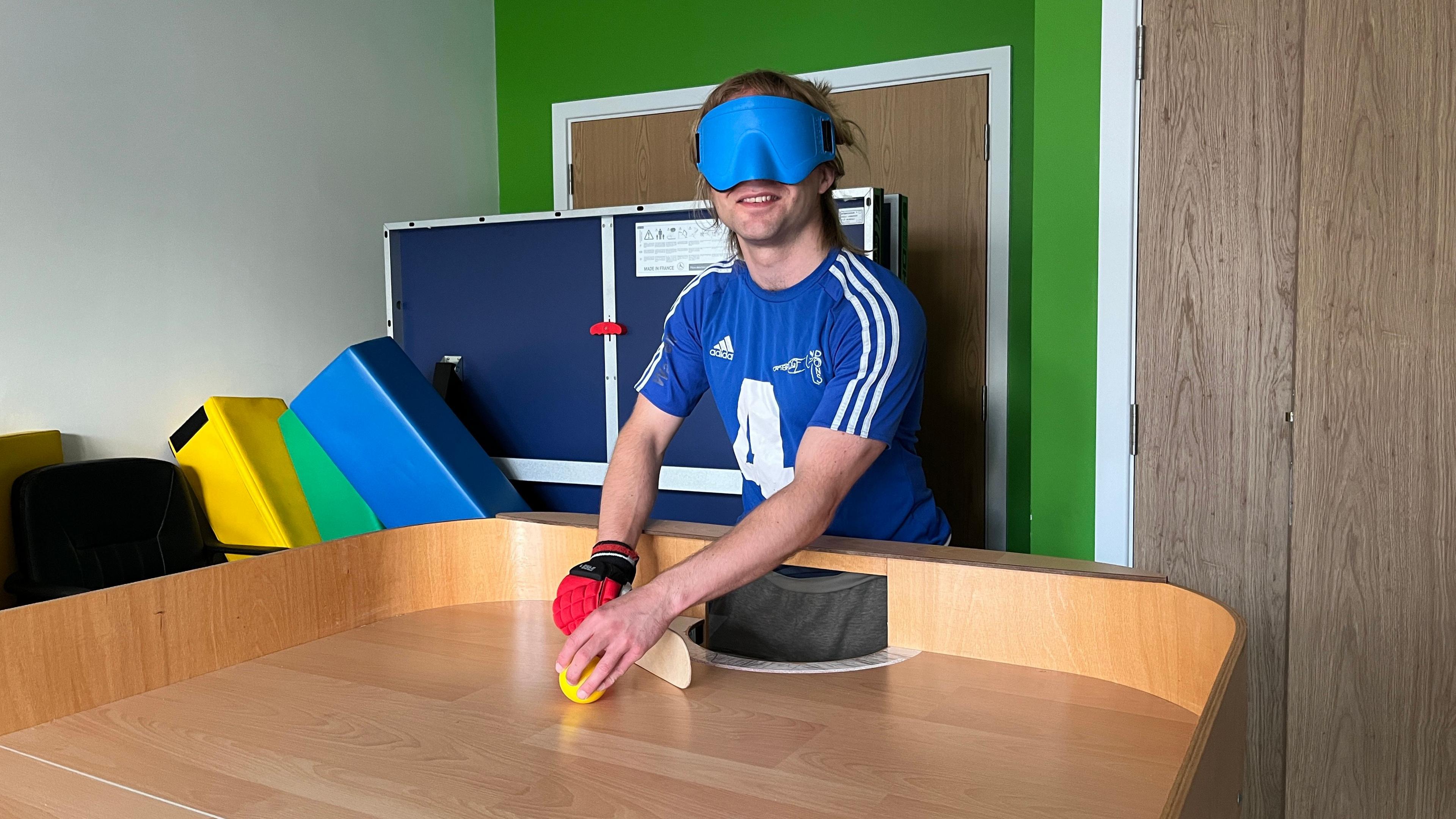 Warren Wilson's face in standing behind the showground table. In one hand he holds a bright yellow ball that is touching the table top. In the other hand he has a small wooden flat bat that his is holding behind the ball - as if he is about to hit it. The table is smooth wood and has large sides around the edges.