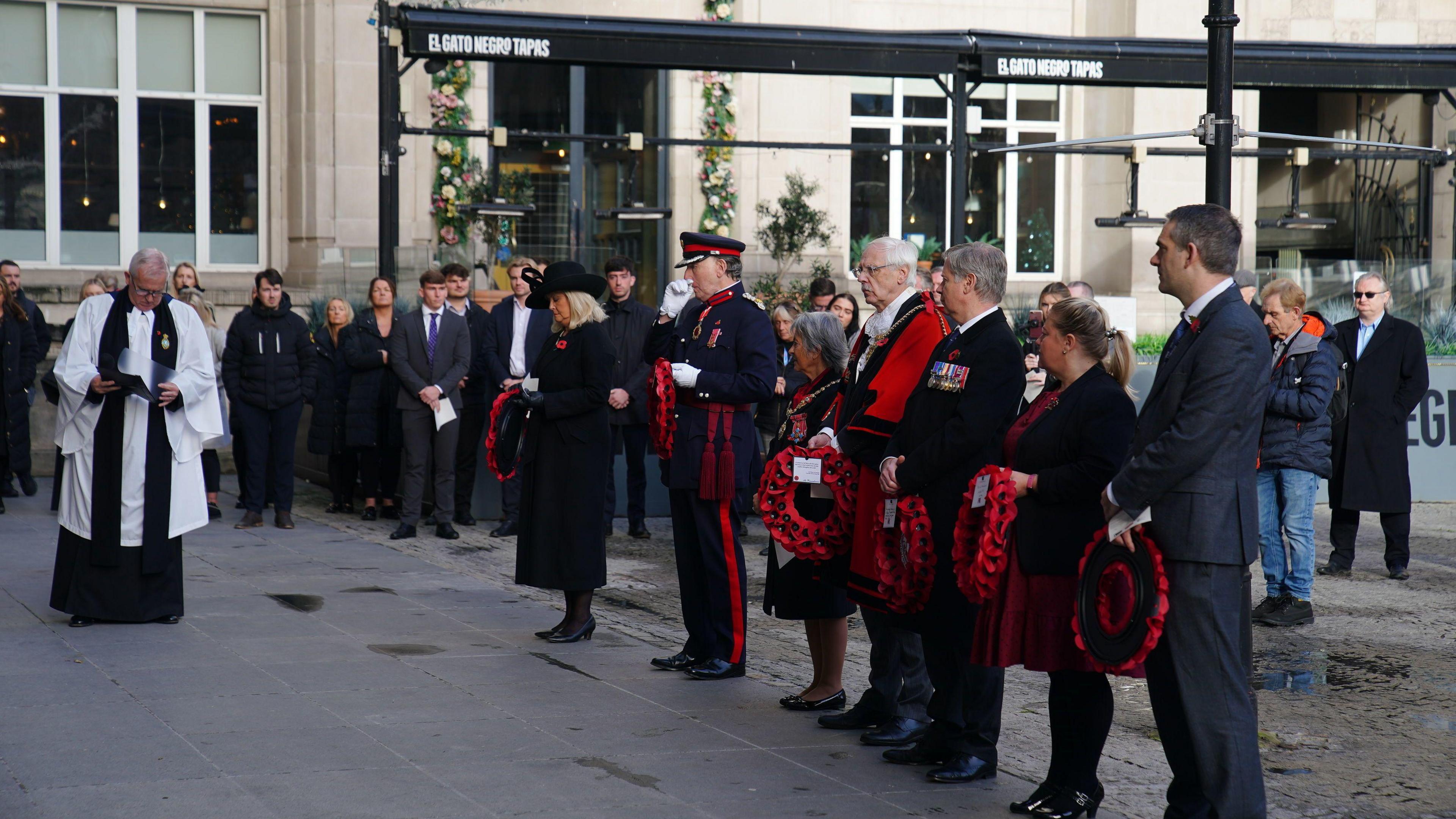 People during a ceremony to mark Armistice Day, at Exchange Flags in Liverpool. A row of people can be seen, some of them with military medals, holding wreaths of poppies - with a man in religious dress standing in front of them and appearing to be reading something. 