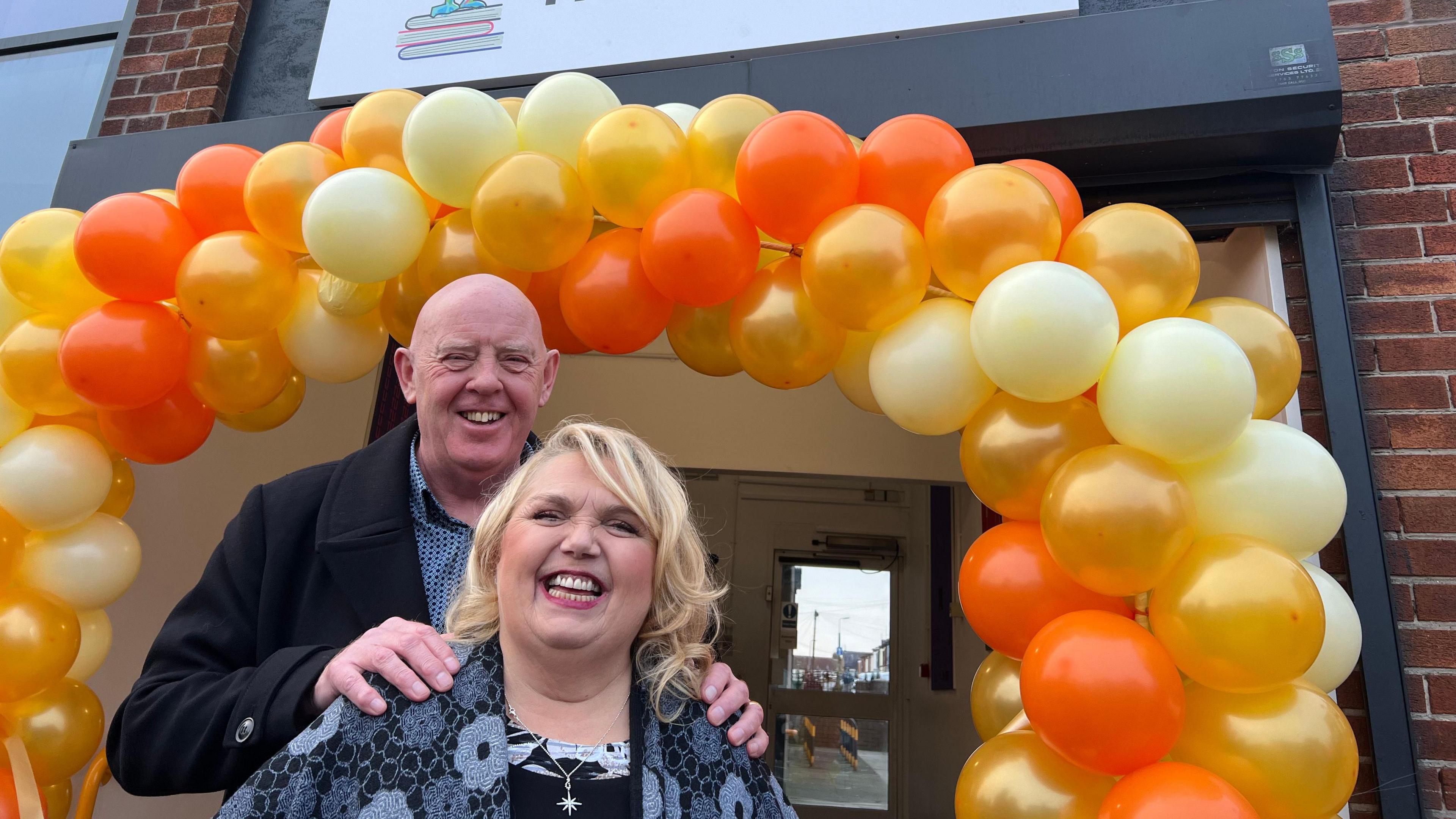 Steve Herron poses with his hands on Sue Herron's shoulders standing in front of an archway made of orange, golden and yellow balloons 