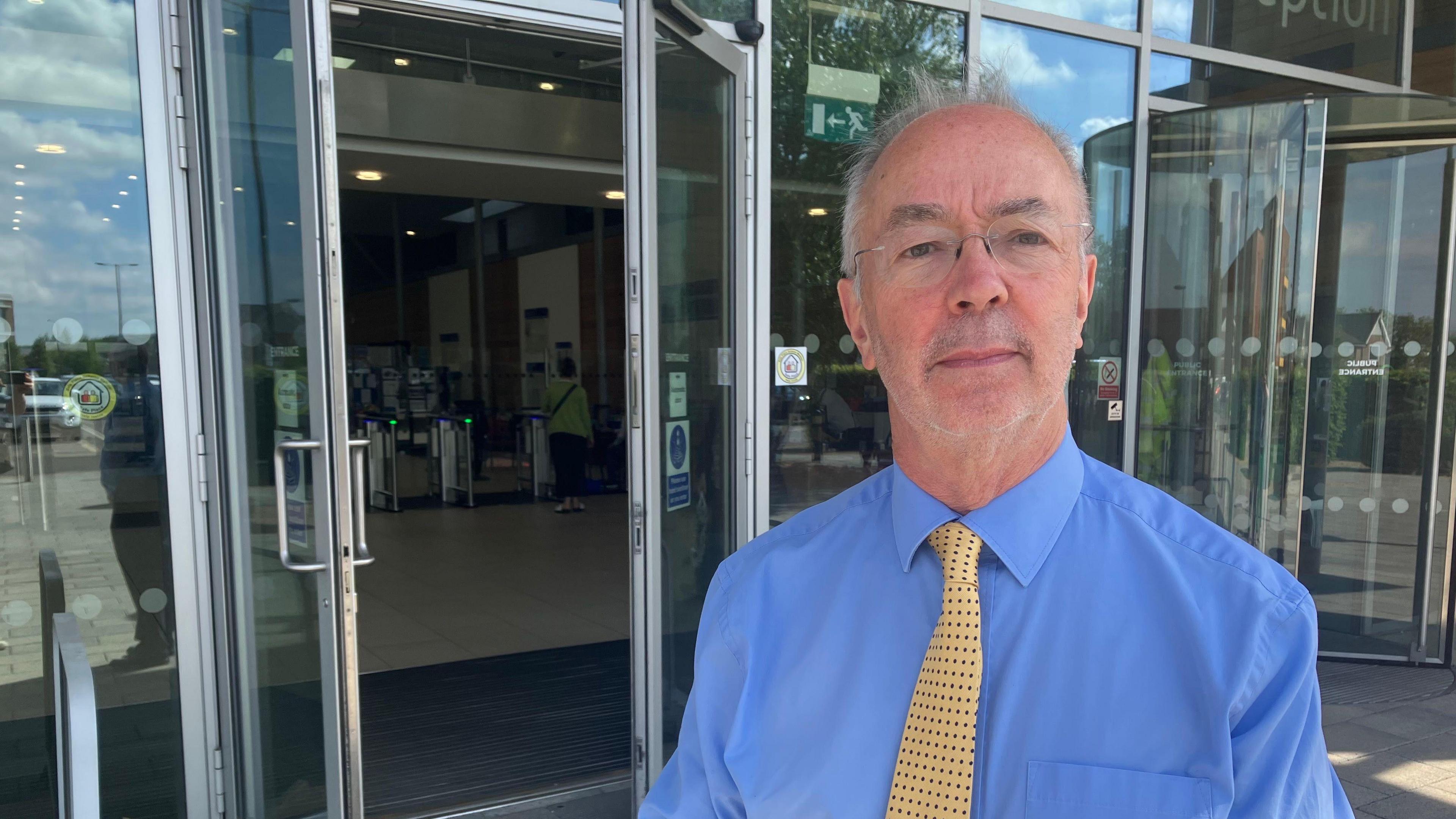 A picture of Buckinghamshire Council leader Martin Tett. He is stood outside the council Gateway building in Aylesbury. He is wearing a blue shirt with a yellow tie with black dots on it.