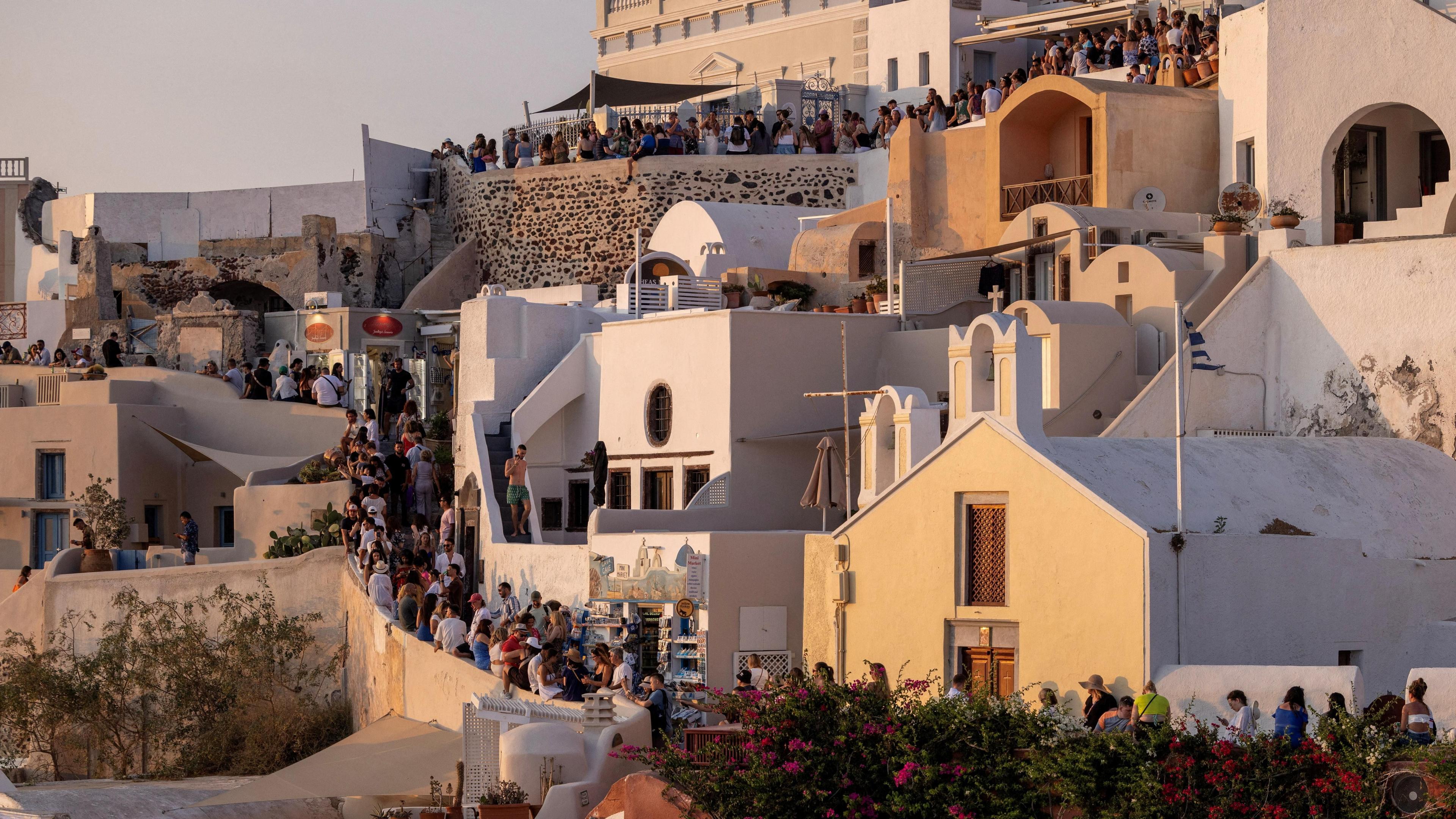 A series of white buildings turn orange as the sunsets on Santorini. Long lines of people can be seen walking to the top of a stair case. Pink bougainvillea is in the foreground. 