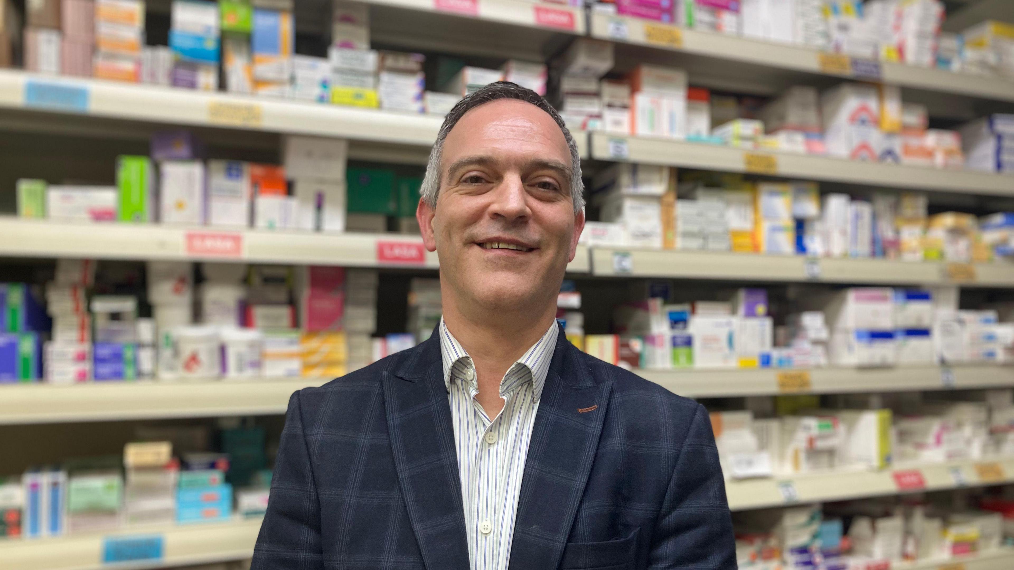 Andrew Hobson looks at the camera wearing a blazer and striped shirt in front of shelves of medicaton