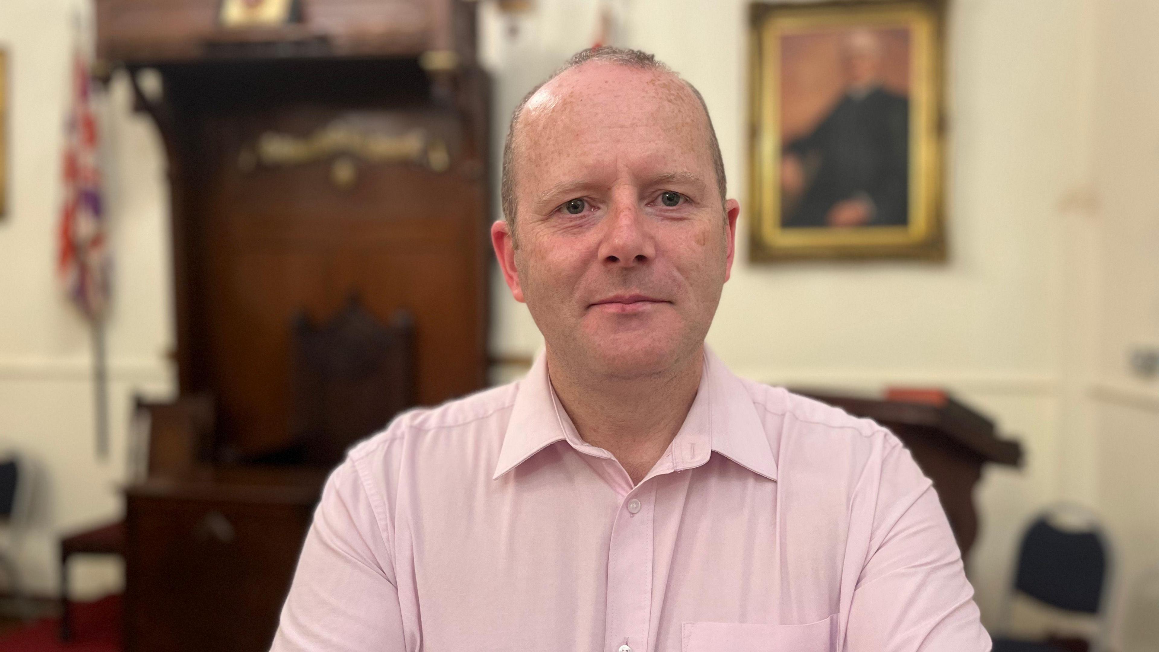 A balding man with grey hair, wearing a pink shirt sat in an office - the background is blurred but a framed image, wooden furniture and union flag are visible