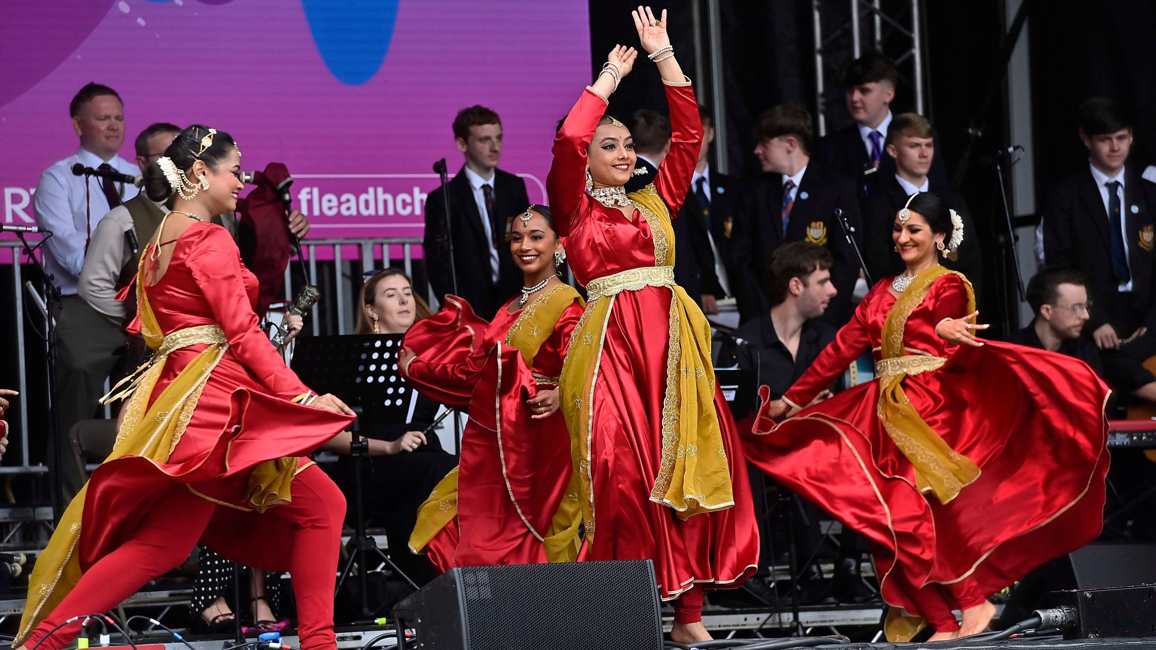 Four women standing on a stage wearing red dresses. Each woman has a gold-coloured piece of fabric adorned. They appear to be dancing. In the background, people stands in suits. 