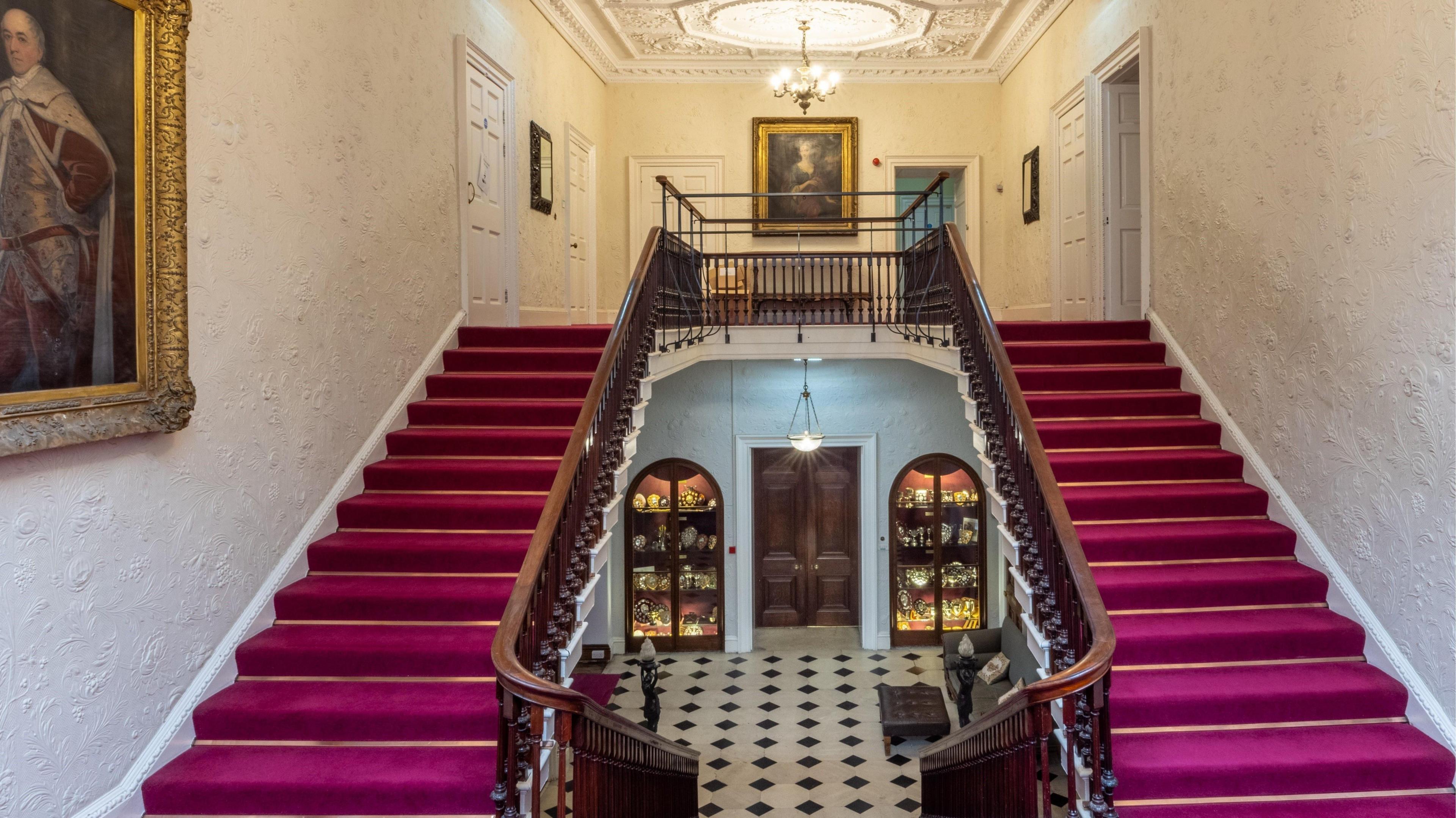 A shot of a double staircase and hallway, with red carpets and white walls decorated with portraits.