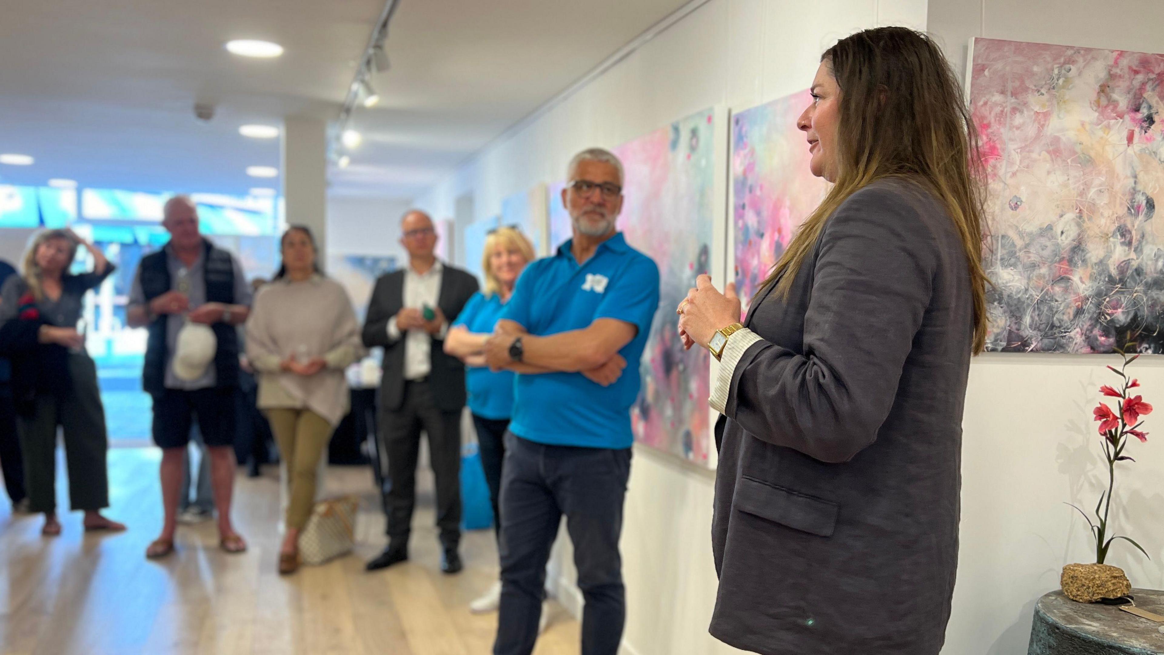 A crowd looks at Emilie who is giving a speech as they stand in the gallery surrounded by her art