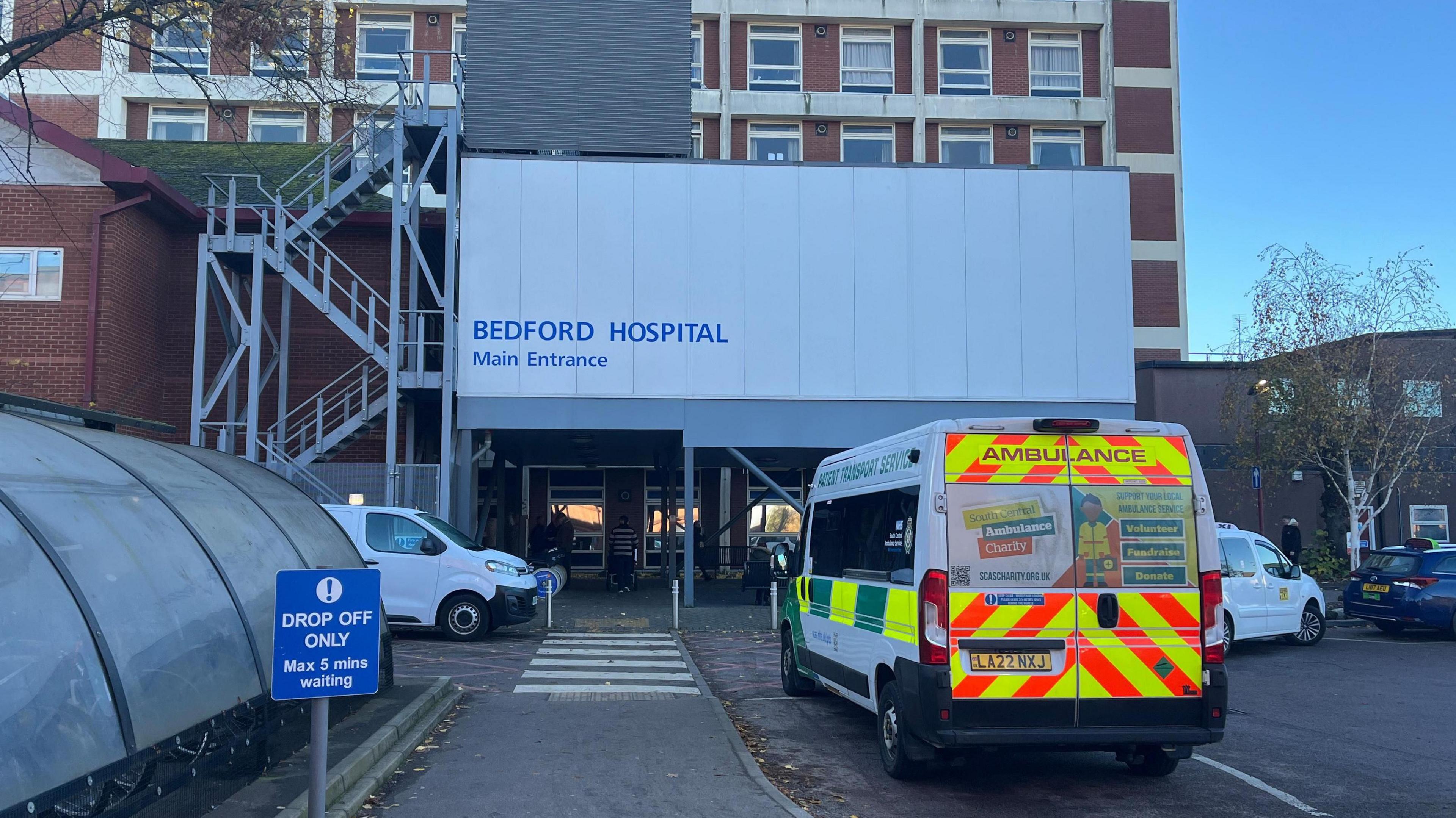 The main entrance of Bedford Hospital with an ambulance outside parked by a drop off zone. There is a pedestrian crossing leading to the entrance.