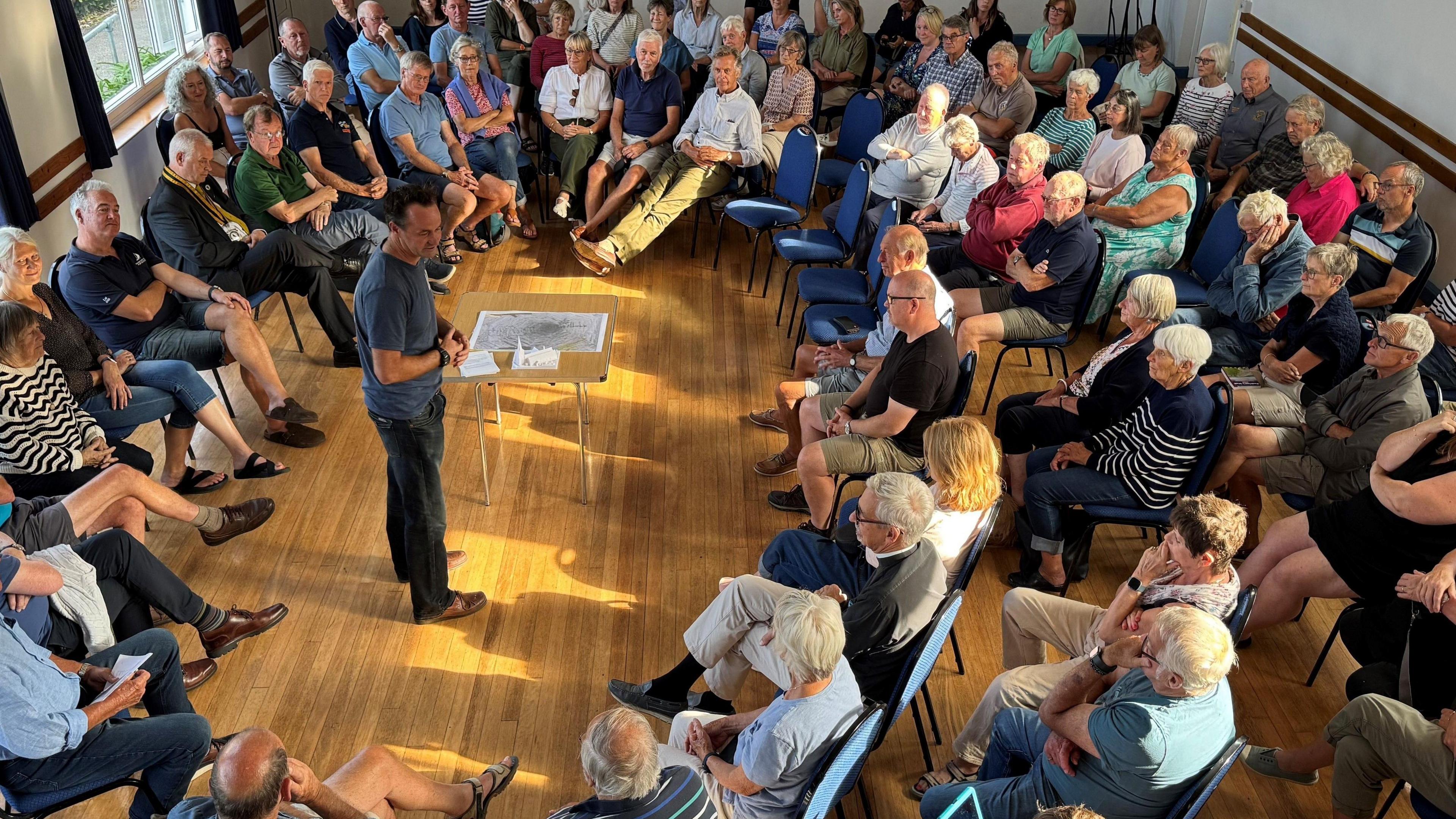 There is a packed room of people sitting on chairs while a man stands in the middle of the room addressing the crowd
