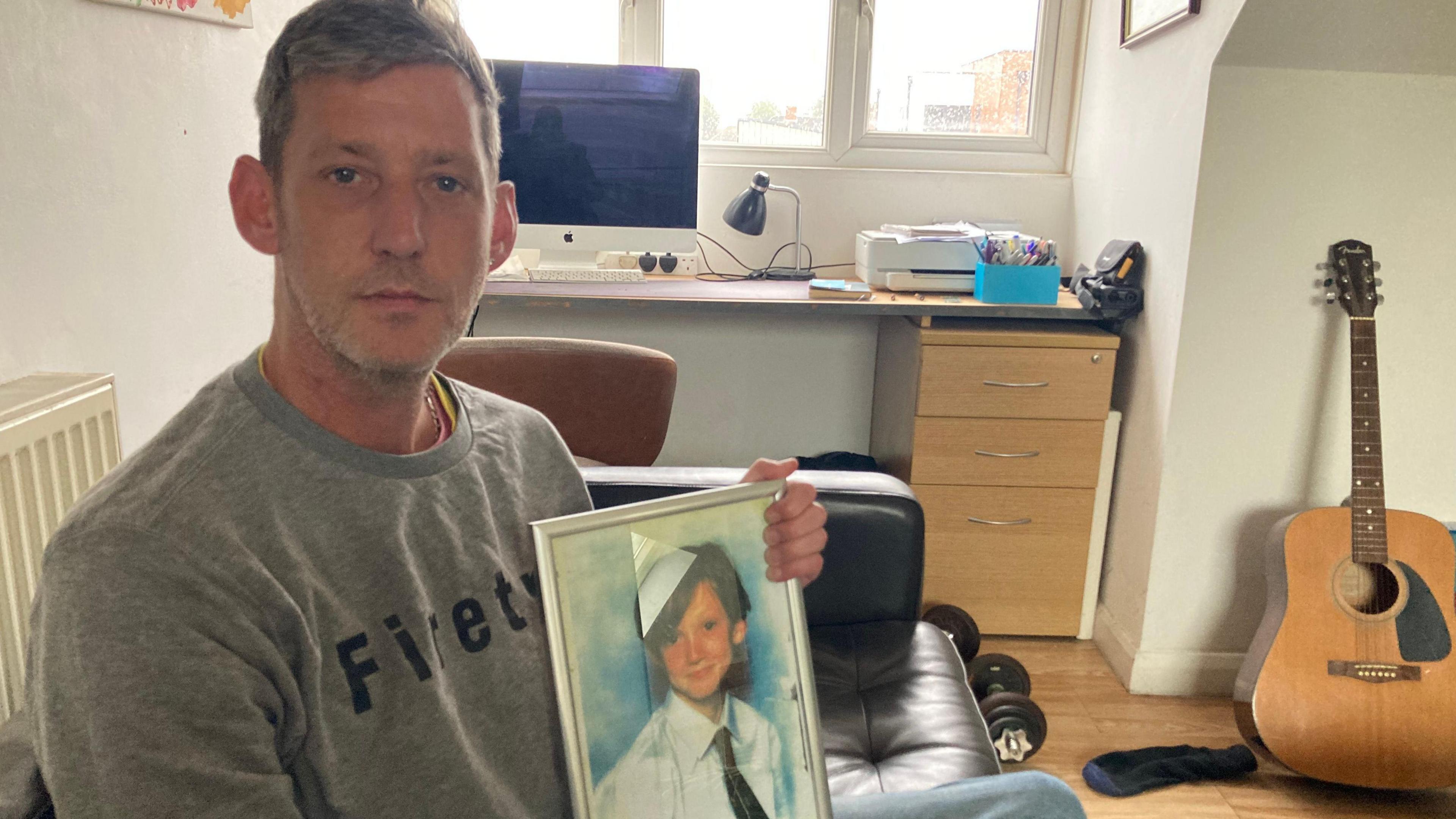 Man with short mousy hair wearing a grey jumper is sat on a sofa by a radiator in front of a window and computer holding a picture of his brother, a young boy who is wearing a school uniform with white shirt and tie. 