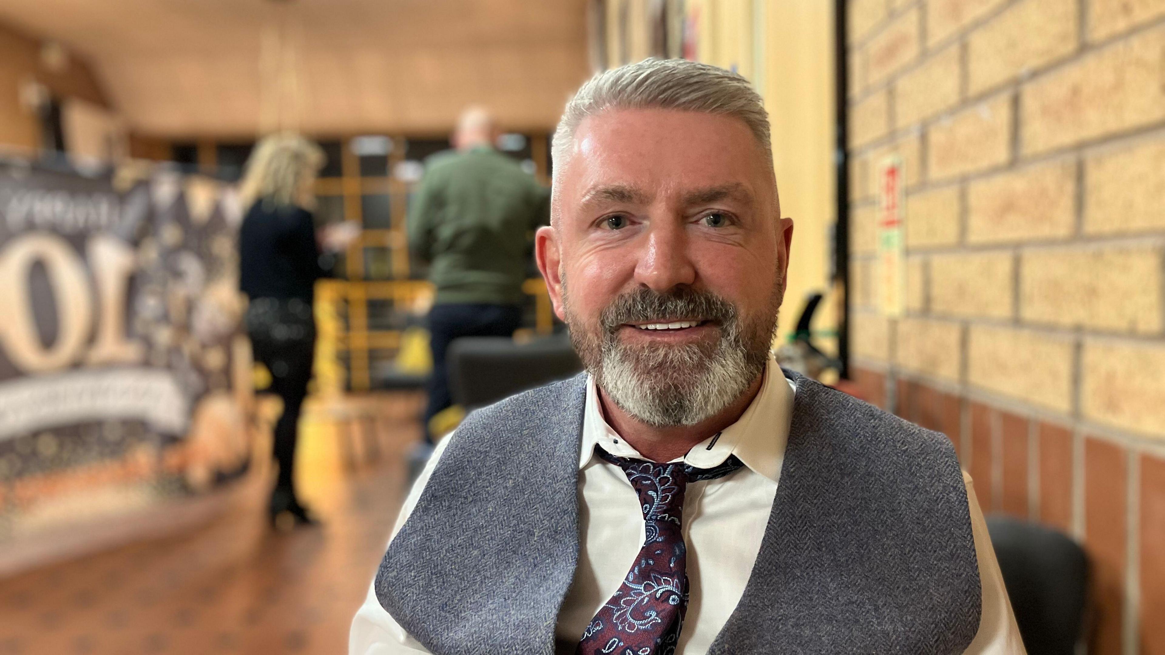 A man with grey hair smiling and looking towards the camera. He has a grey waistcoat on with a whit shirt and a maroon and teal tie.