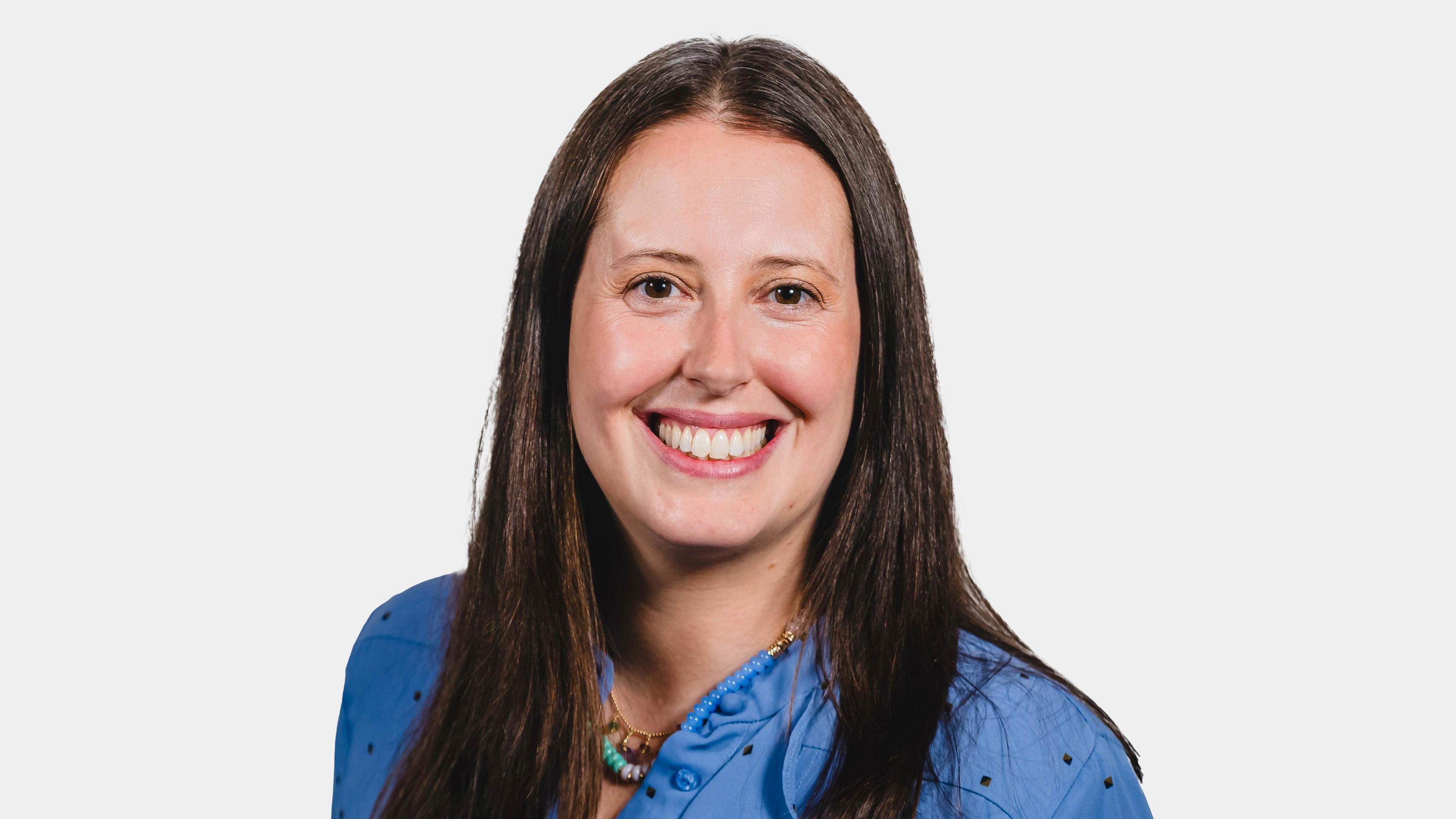 A woman with long dark hair smiles at the camera. She is standing in front of a plain background and is wearing a blue shirt.